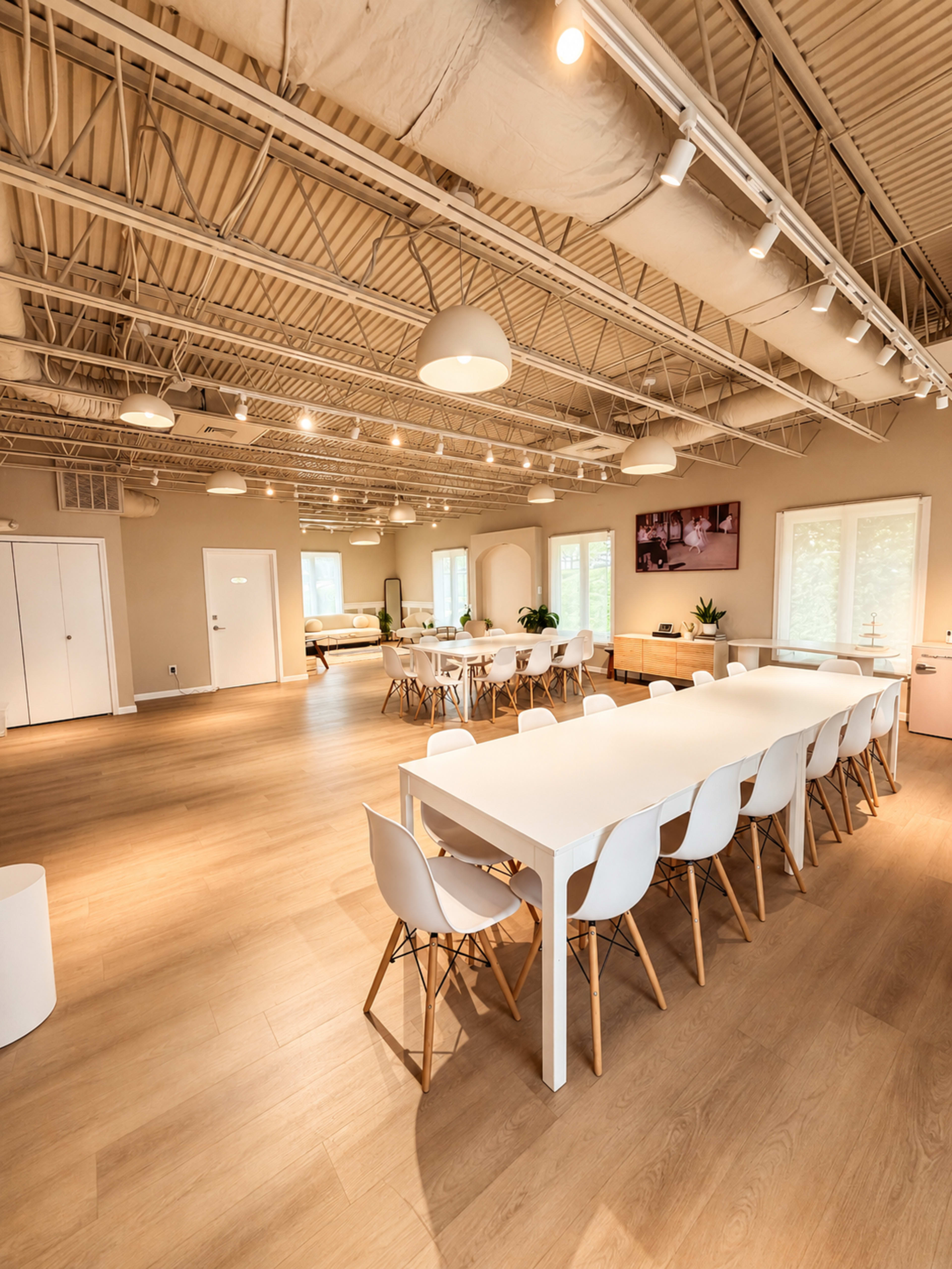 The image shows a spacious, well-lit modern office featuring a long white table surrounded by white chairs, with wooden floors and large windows.