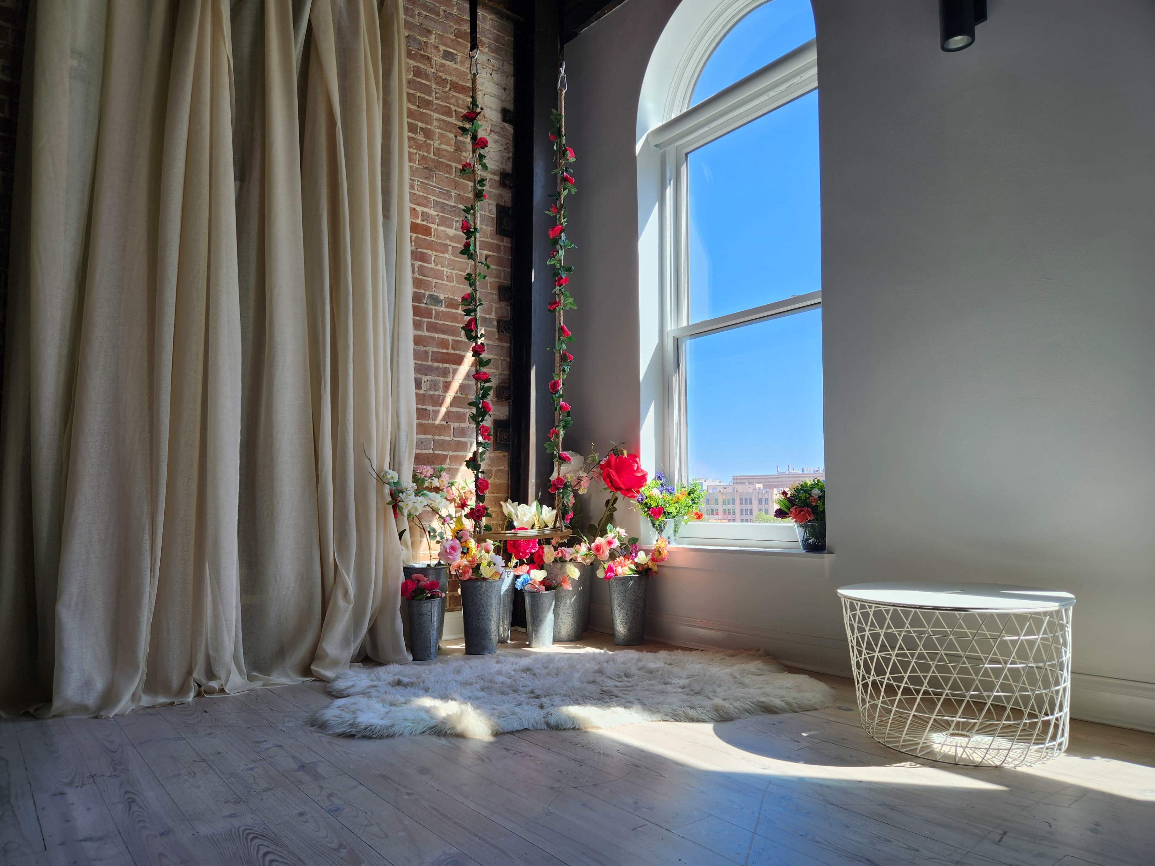 A sunlit corner of a room features a window adorned with flowers in metal pots, beside a white basket and draped curtains against a brick wall.