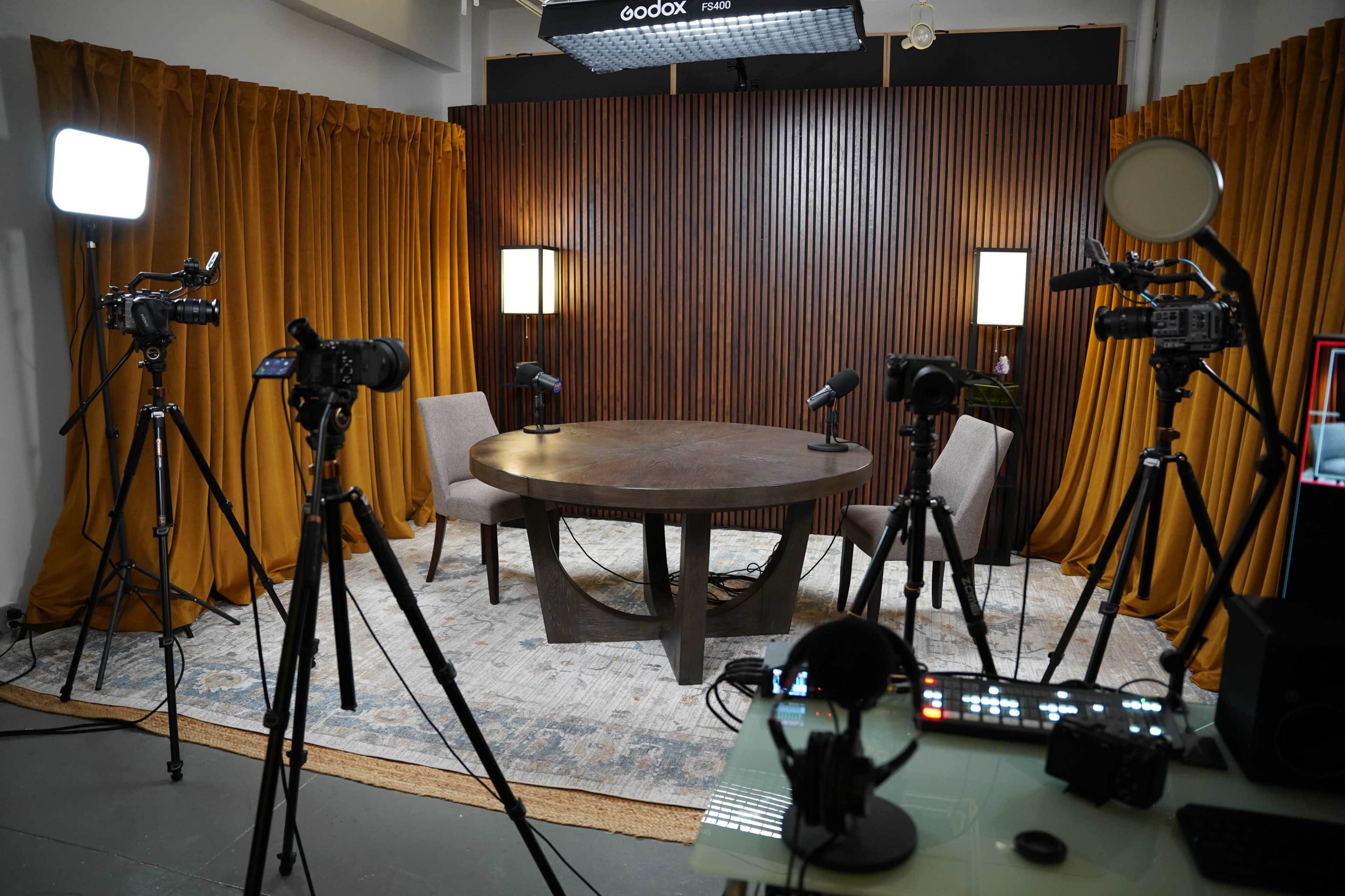 The image shows a recording studio setup featuring a round table surrounded by two chairs, with several cameras on tripods and lighting equipment in the background.