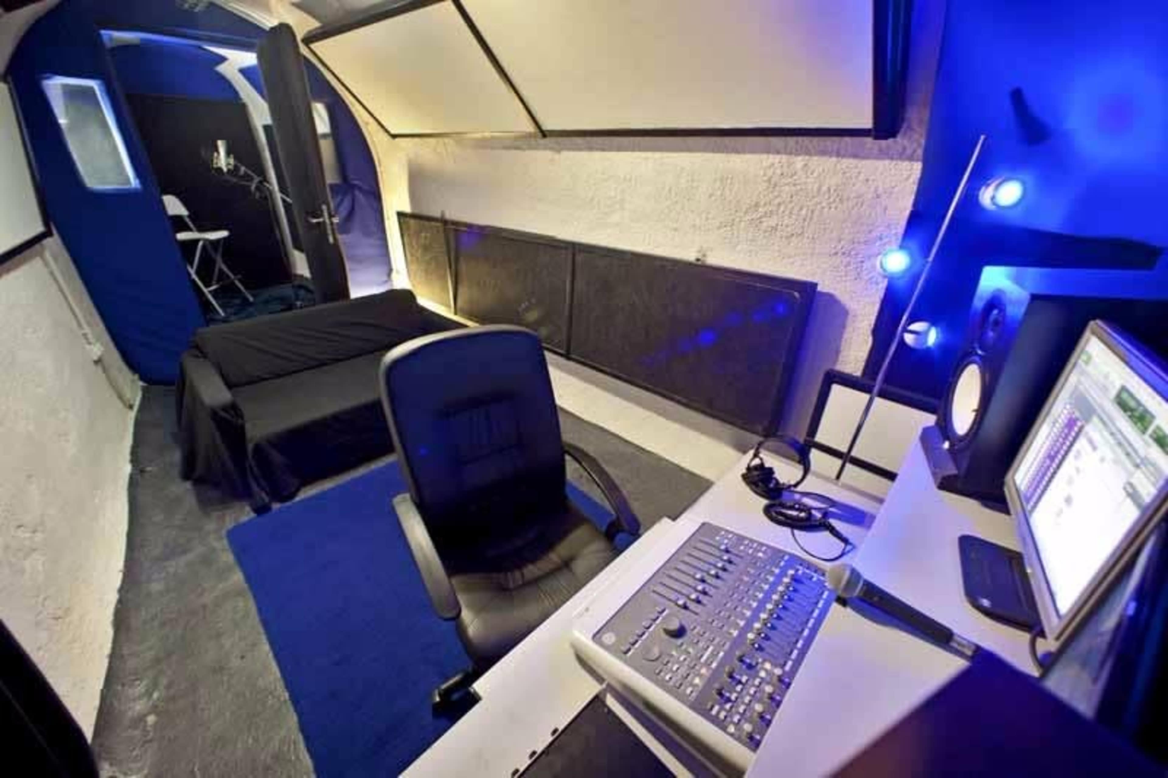 The image shows a compact studio space with a desk and computer setup, a black bed, and a chair positioned on a blue rug.