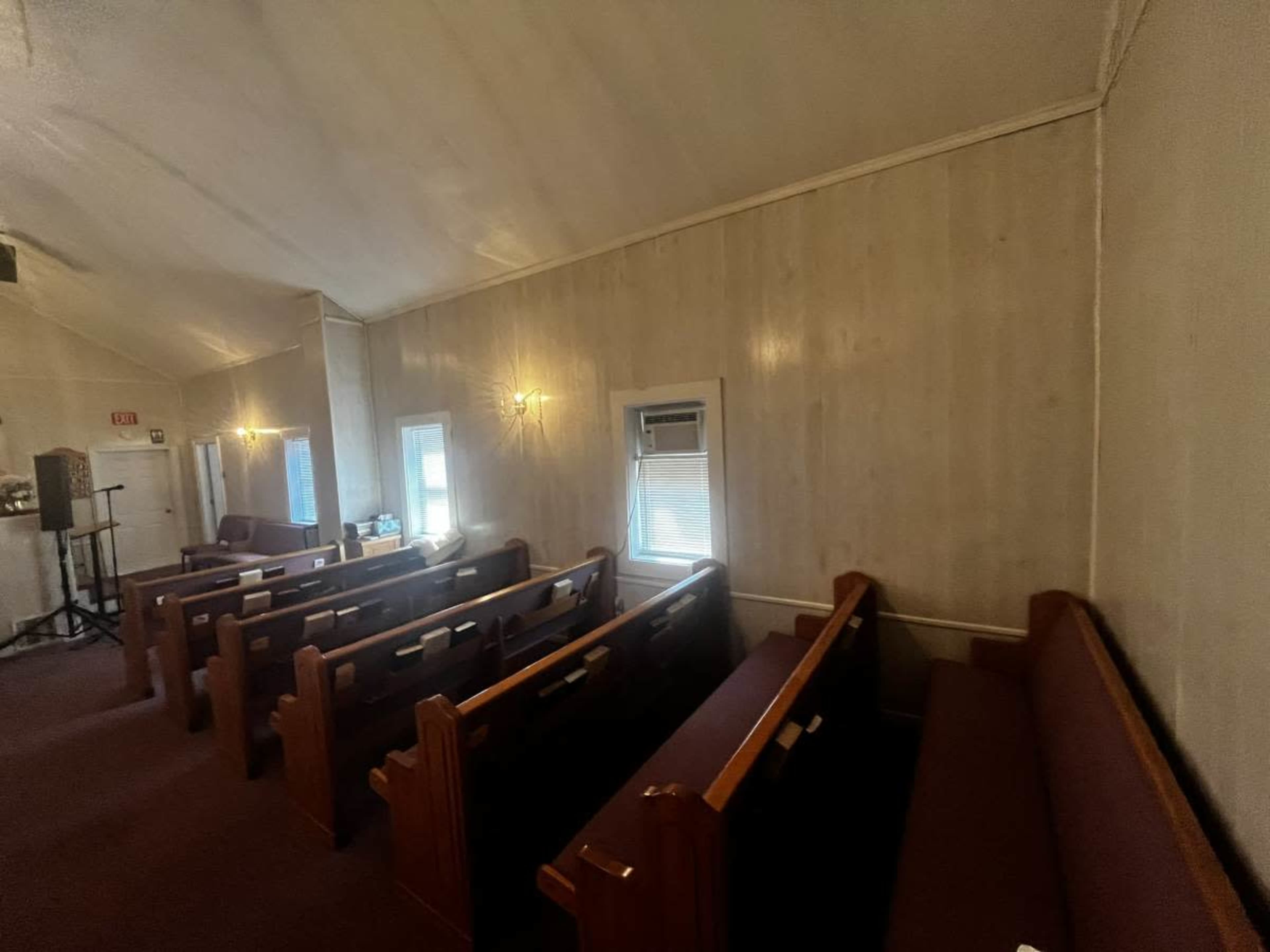 The image shows the interior of a small church with wooden pews arranged along a carpeted aisle and windows providing natural light.