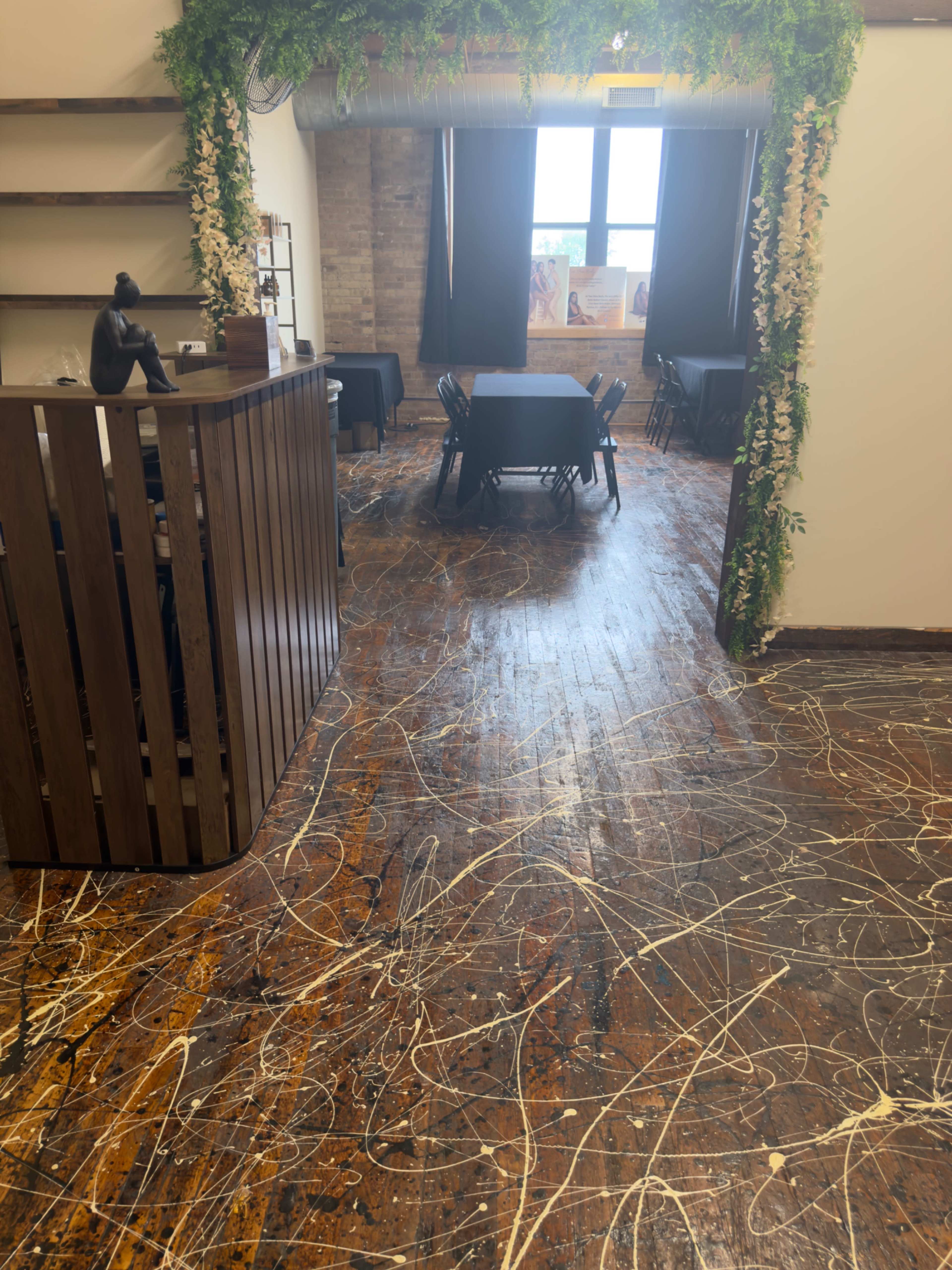 The image shows a room with wooden floors featuring scattered paint splatters, a reception area with a wooden counter on the left, and tables set up for dining in the background.