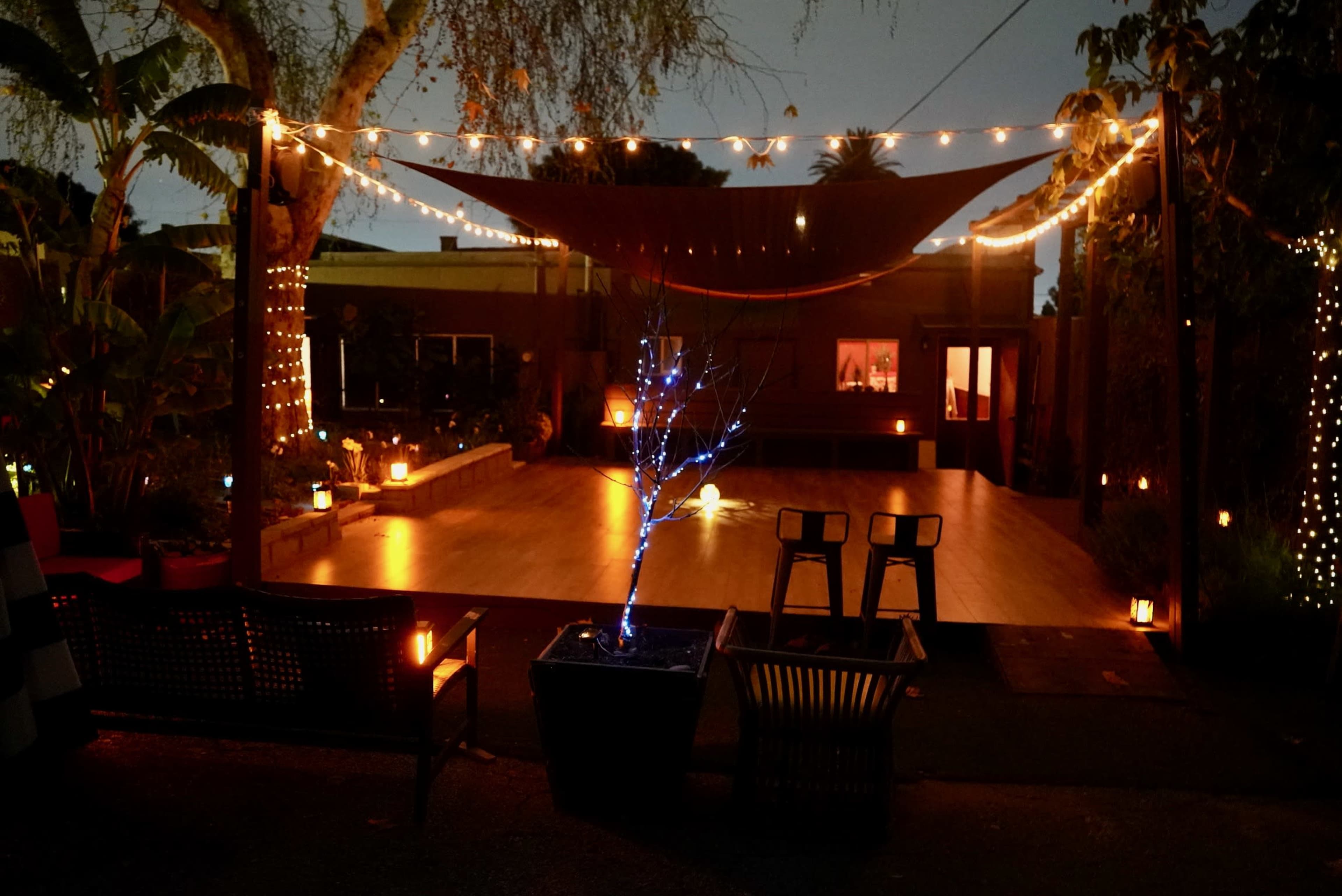 A well-lit outdoor space features a wooden deck with string lights, a small tree with blue lights, and two chairs facing the inviting interior of a building.