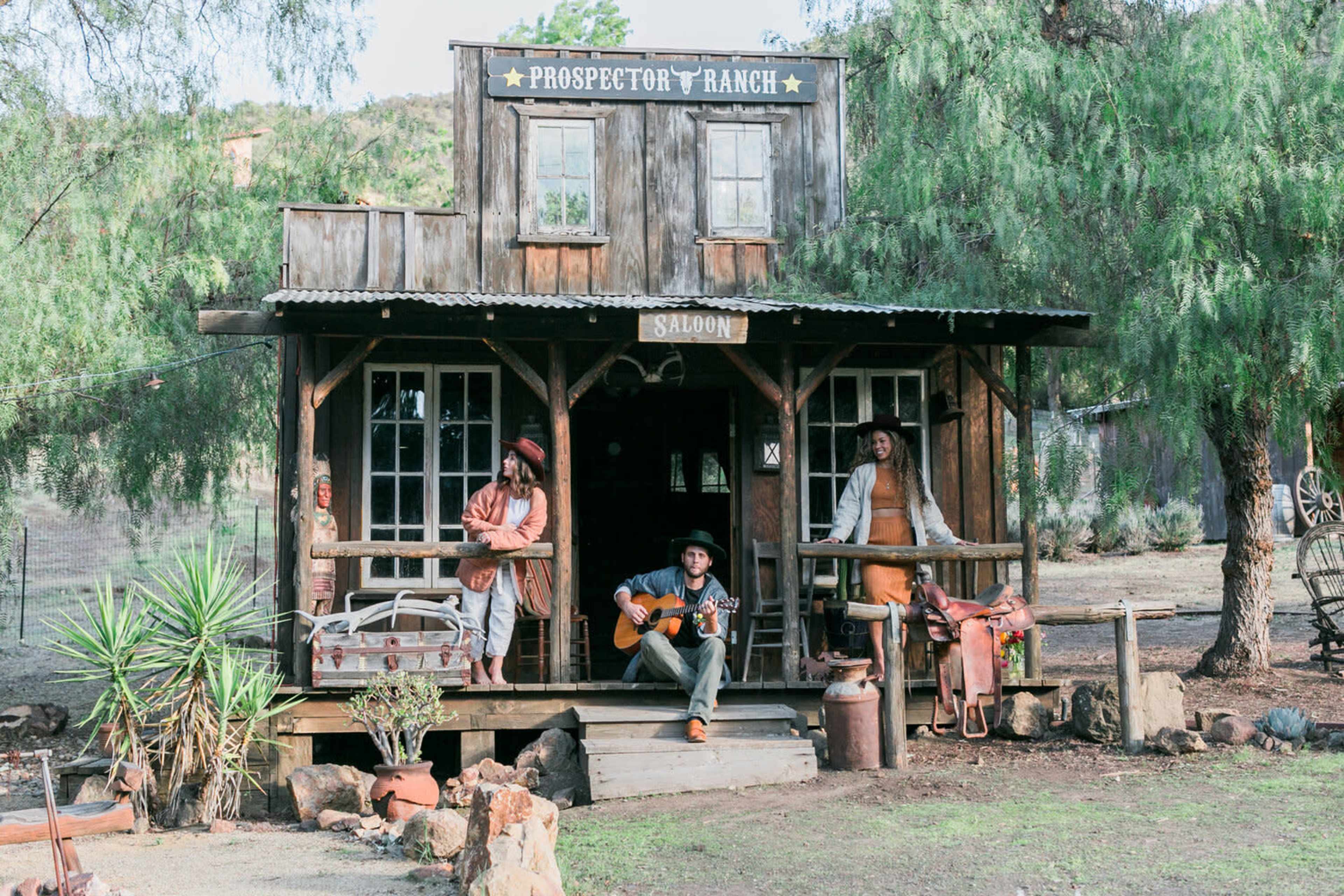 A cowboy sits on the steps of a wooden saloon, playing guitar, while two women pose on the porch at Prospector Ranch.