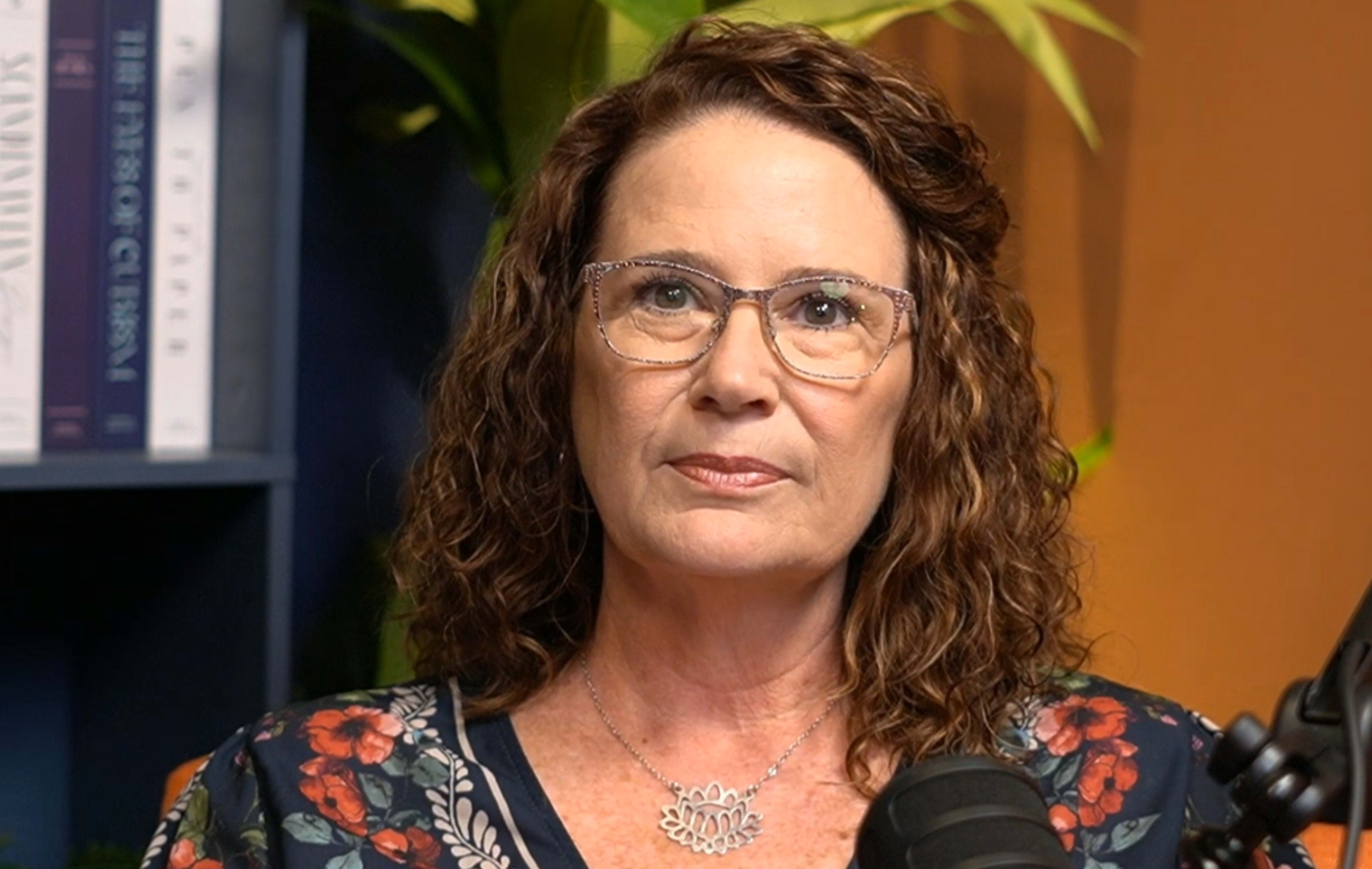 A woman with curly hair and glasses is seated in front of a bookshelf and a plant.