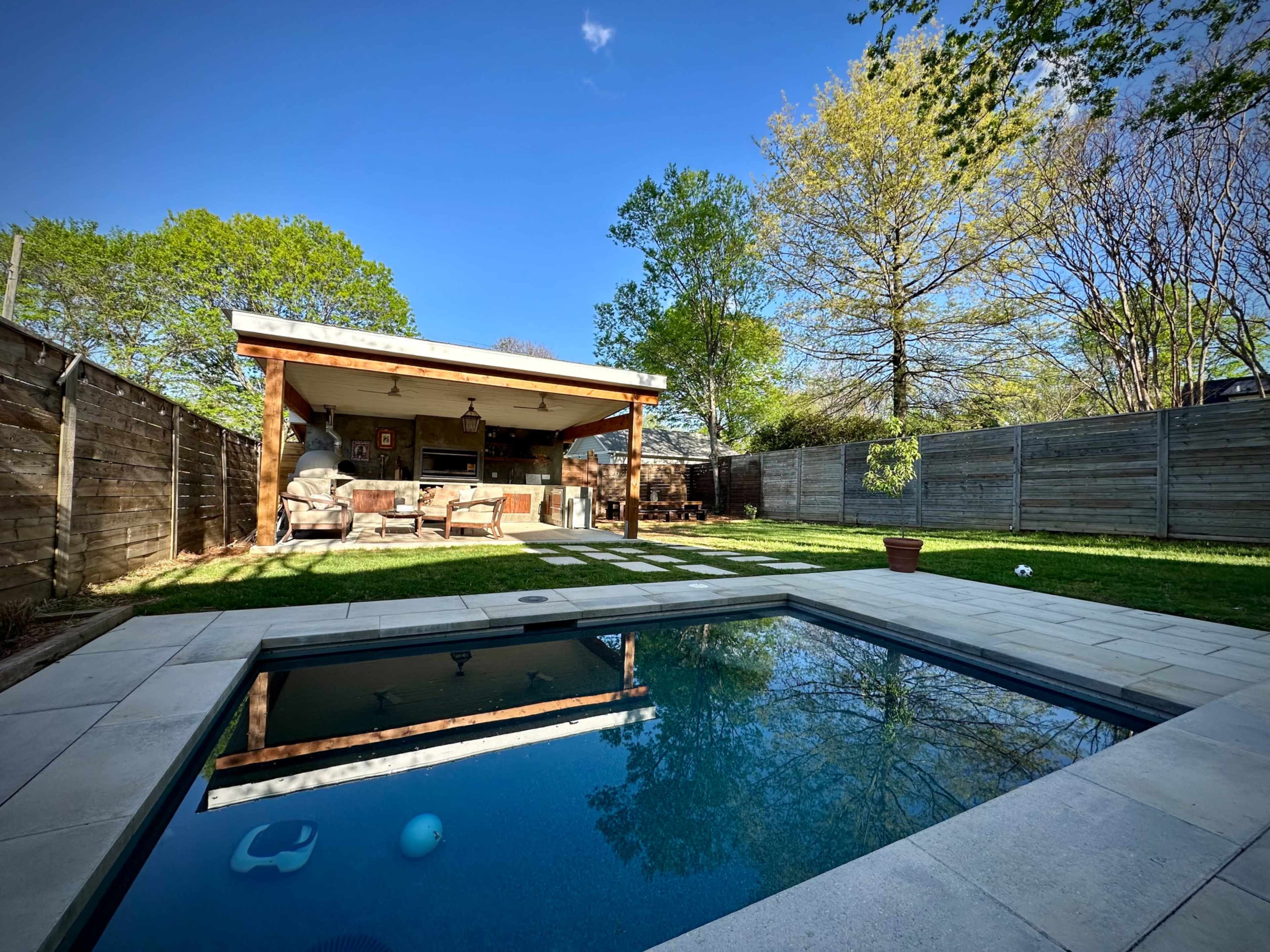 The image shows a backyard with a swimming pool in the foreground and a covered patio area with seating in the background, surrounded by trees and a wooden fence.