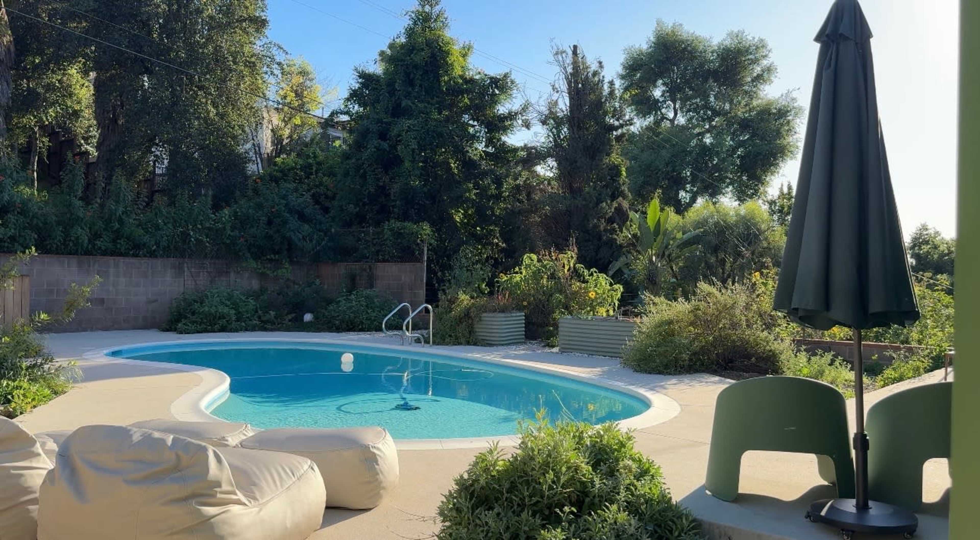 The scene features a swimming pool surrounded by lush greenery and a few lounge chairs and an umbrella nearby.