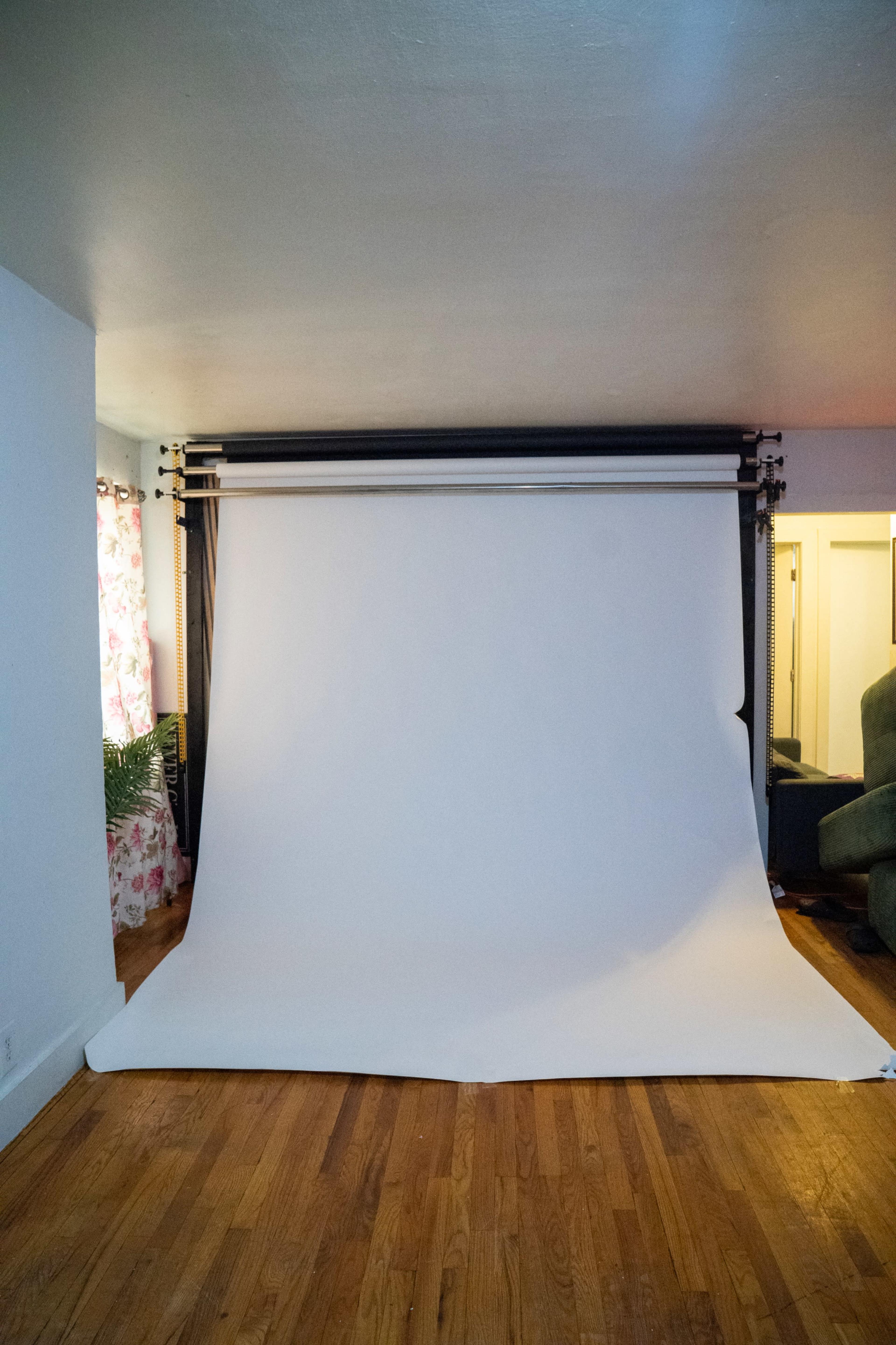 The image shows a bright, uncluttered room with a white backdrop set up on a support system and light hardwood flooring.