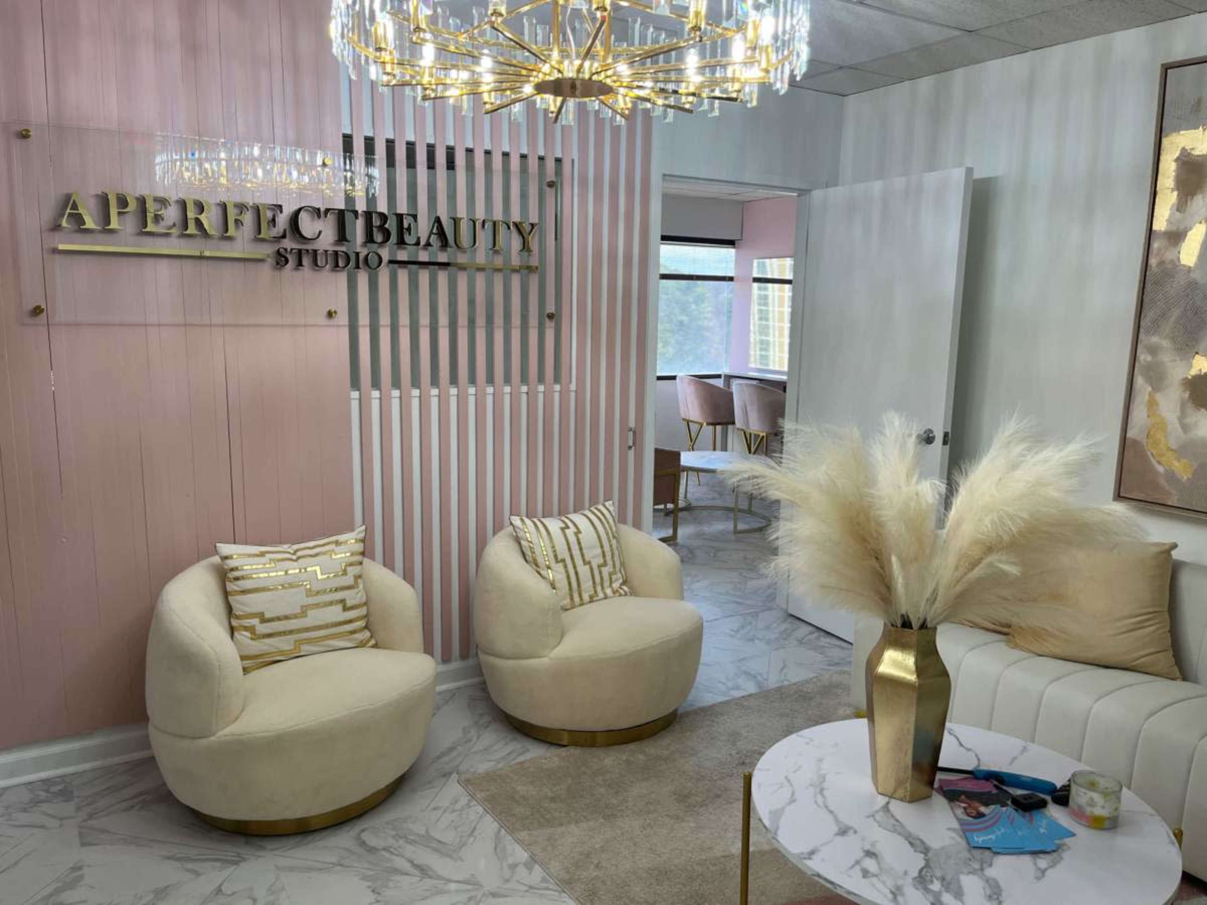 A modern beauty studio reception area with cream-colored chairs, a round coffee table, and a prominent sign reading "APERFECTBEAUTY STUDIO" mounted on a decorative wall.