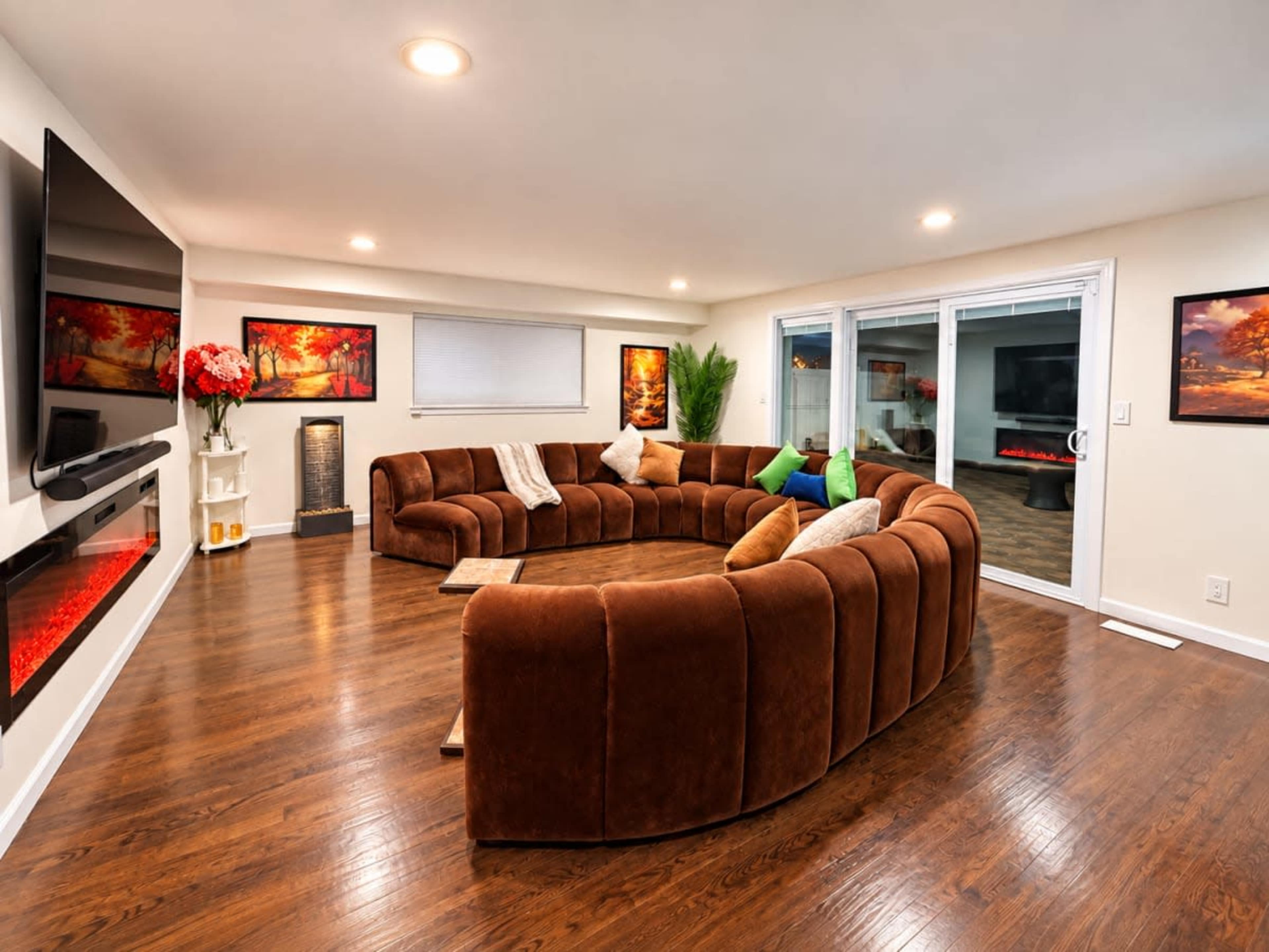 The image shows a modern living room featuring a large, curved brown sofa arranged around a central circular coffee table, with a flat-screen TV mounted on the wall and sliding glass doors leading to an outdoor area.