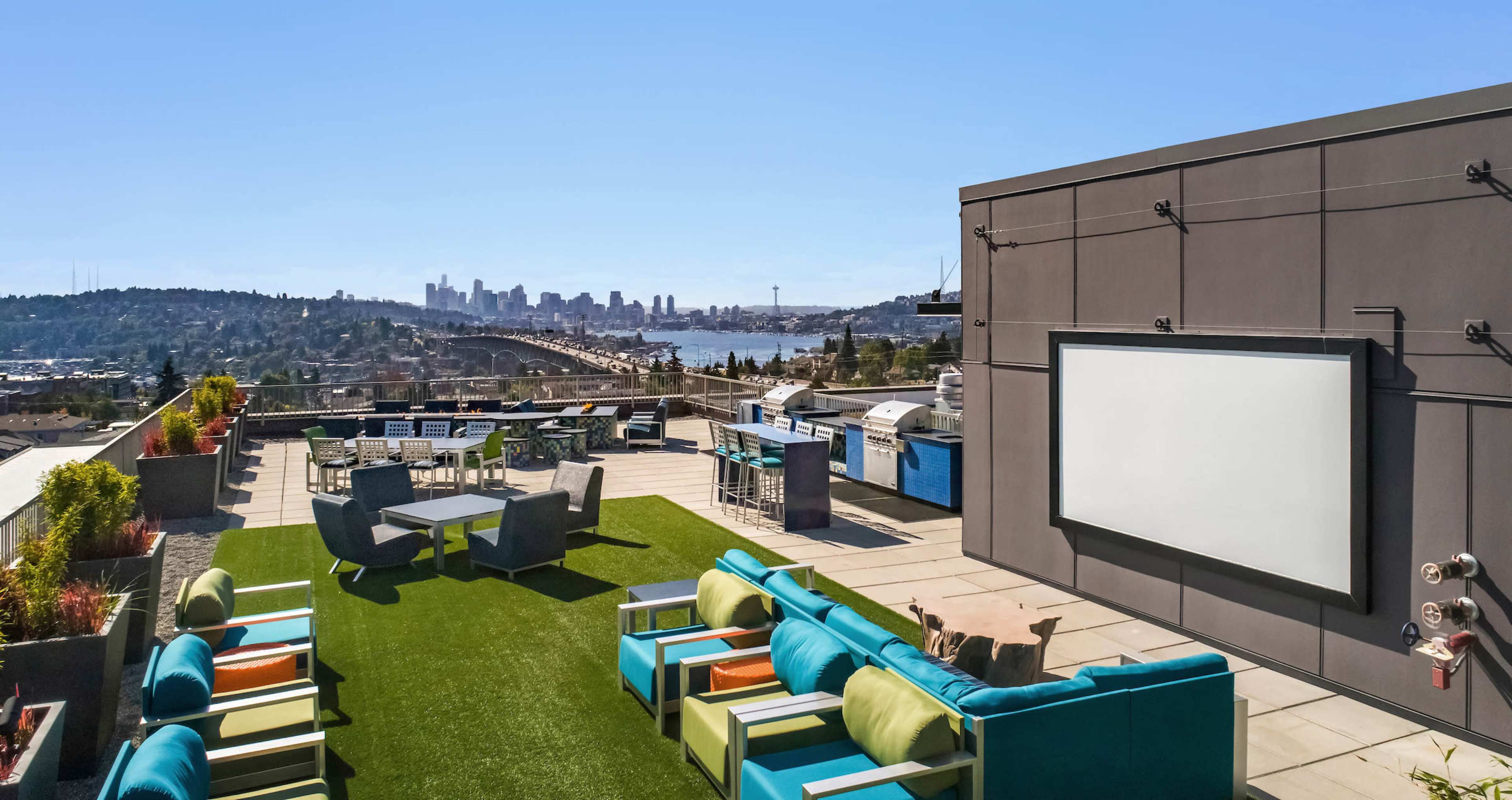 A rooftop terrace features colorful seating and an outdoor screen with a view of a city skyline and a body of water.