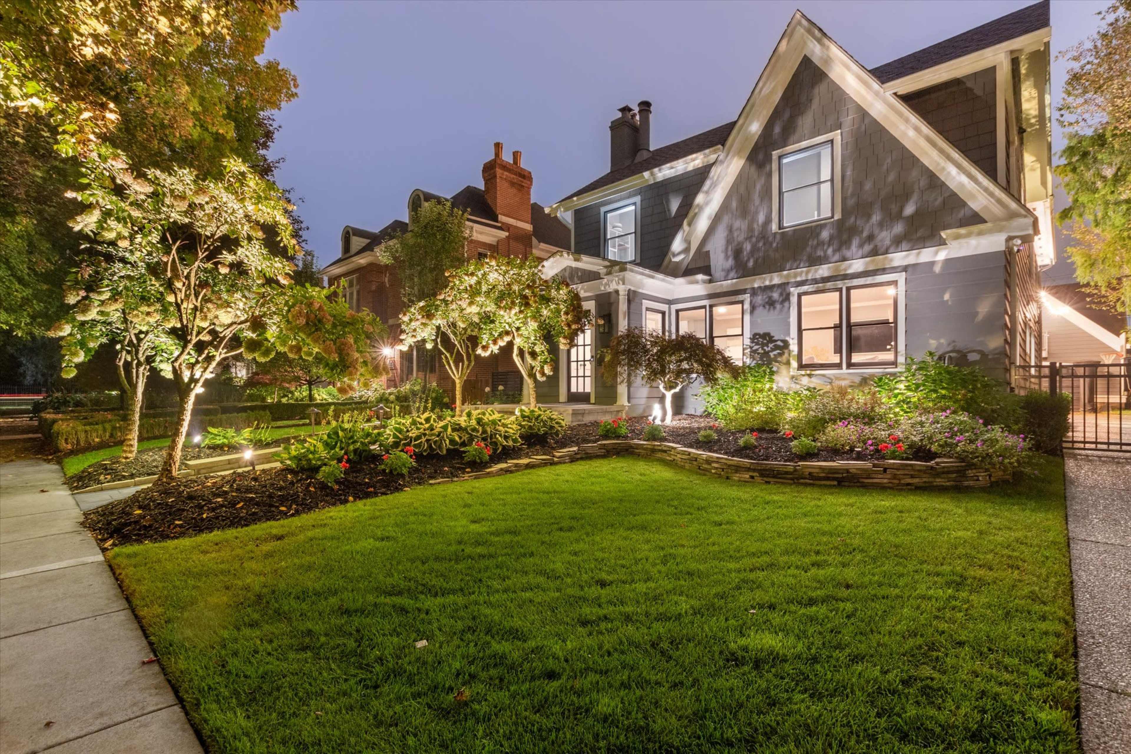 A well-lit, modern house with a manicured lawn and flower beds is seen at dusk.