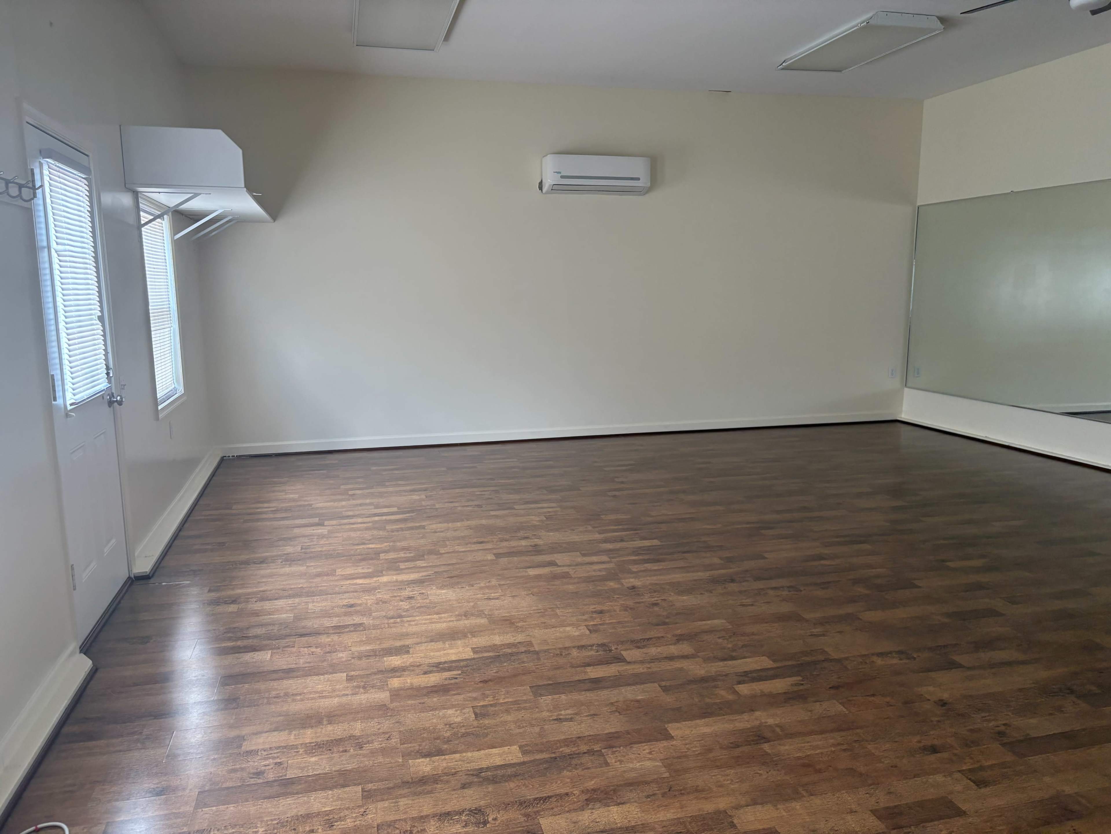 The image shows an empty room with hardwood floors, a large mirror on one wall, and two windows allowing natural light.