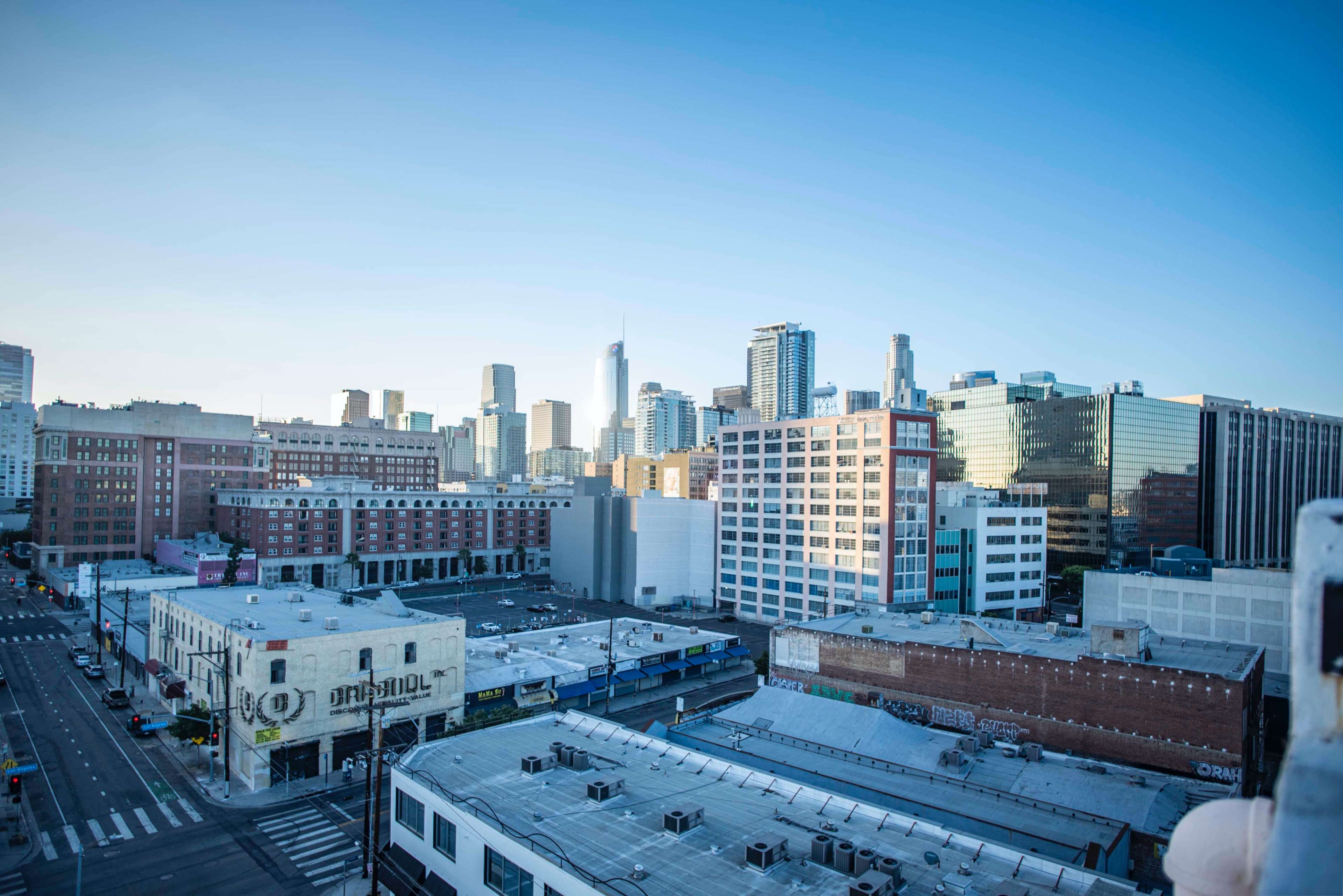 The image shows a skyline of tall buildings in a city, with a clear blue sky above and a street lined with low-rise structures in the foreground.