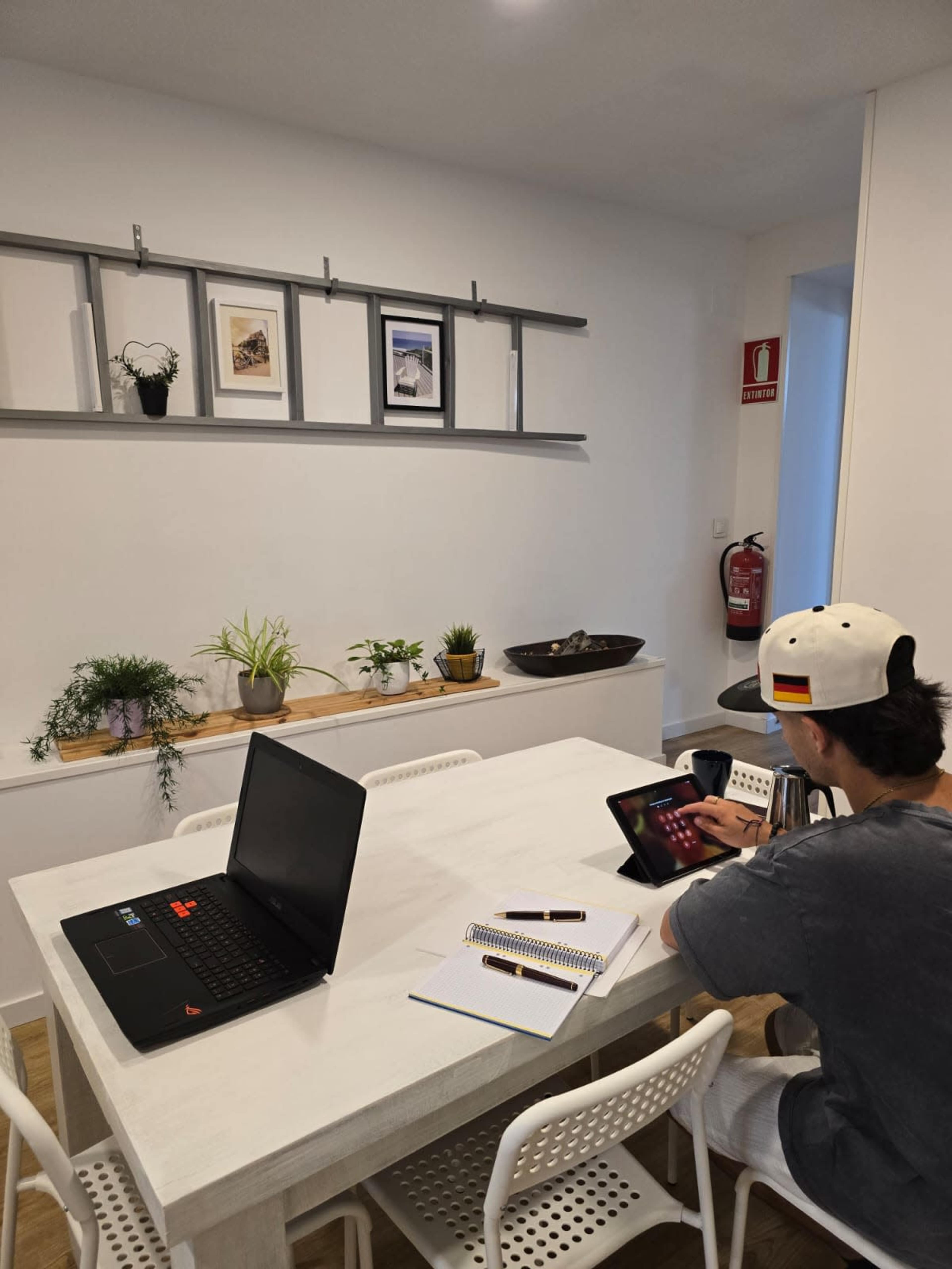 A person sits at a table working on a tablet, with a laptop and notebook nearby in a well-lit room featuring plants and framed pictures on the wall.