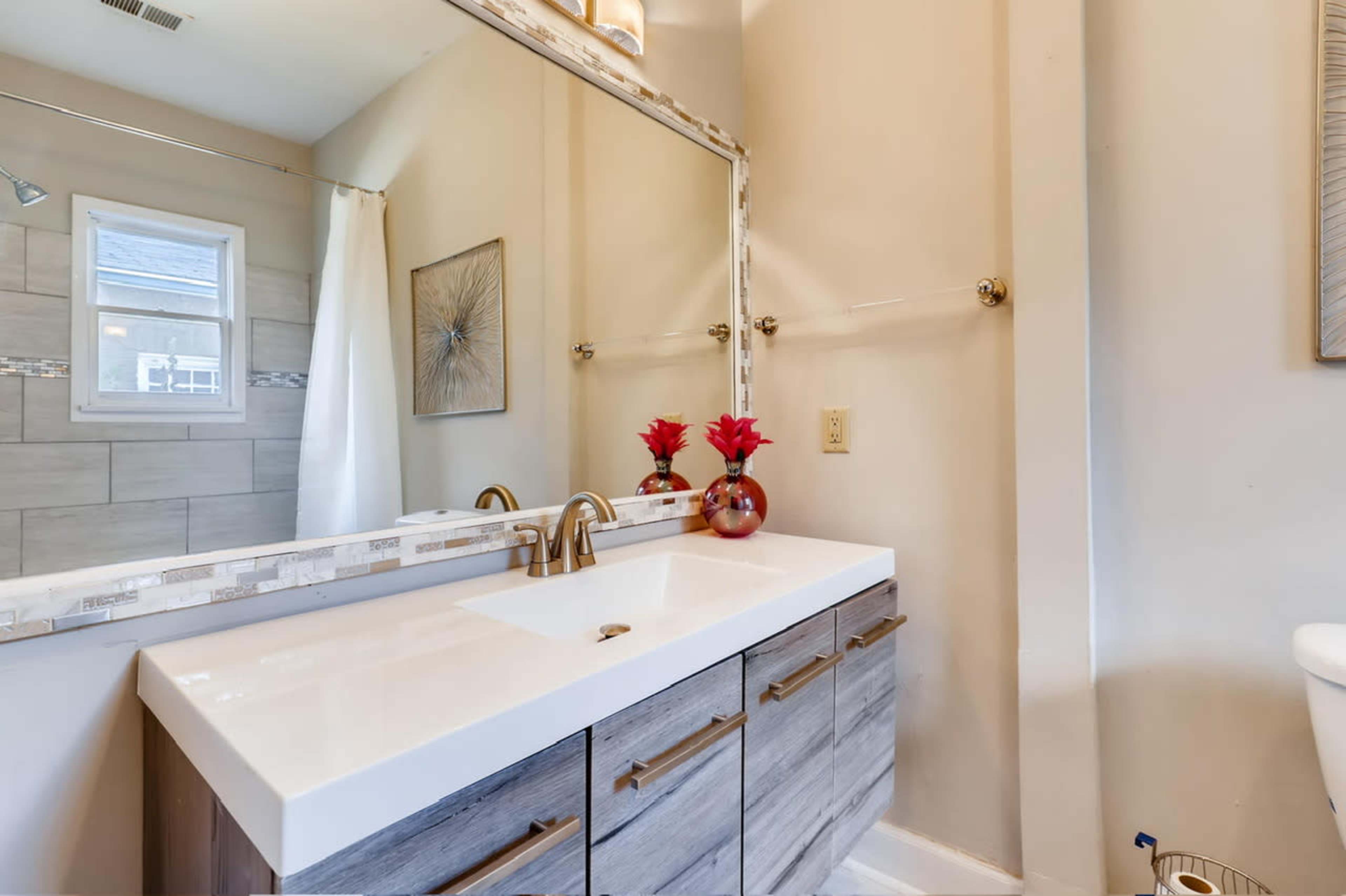 The image depicts a modern bathroom with a double sink vanity featuring a white countertop and wooden drawers, complemented by a large mirror and decorative flowers.