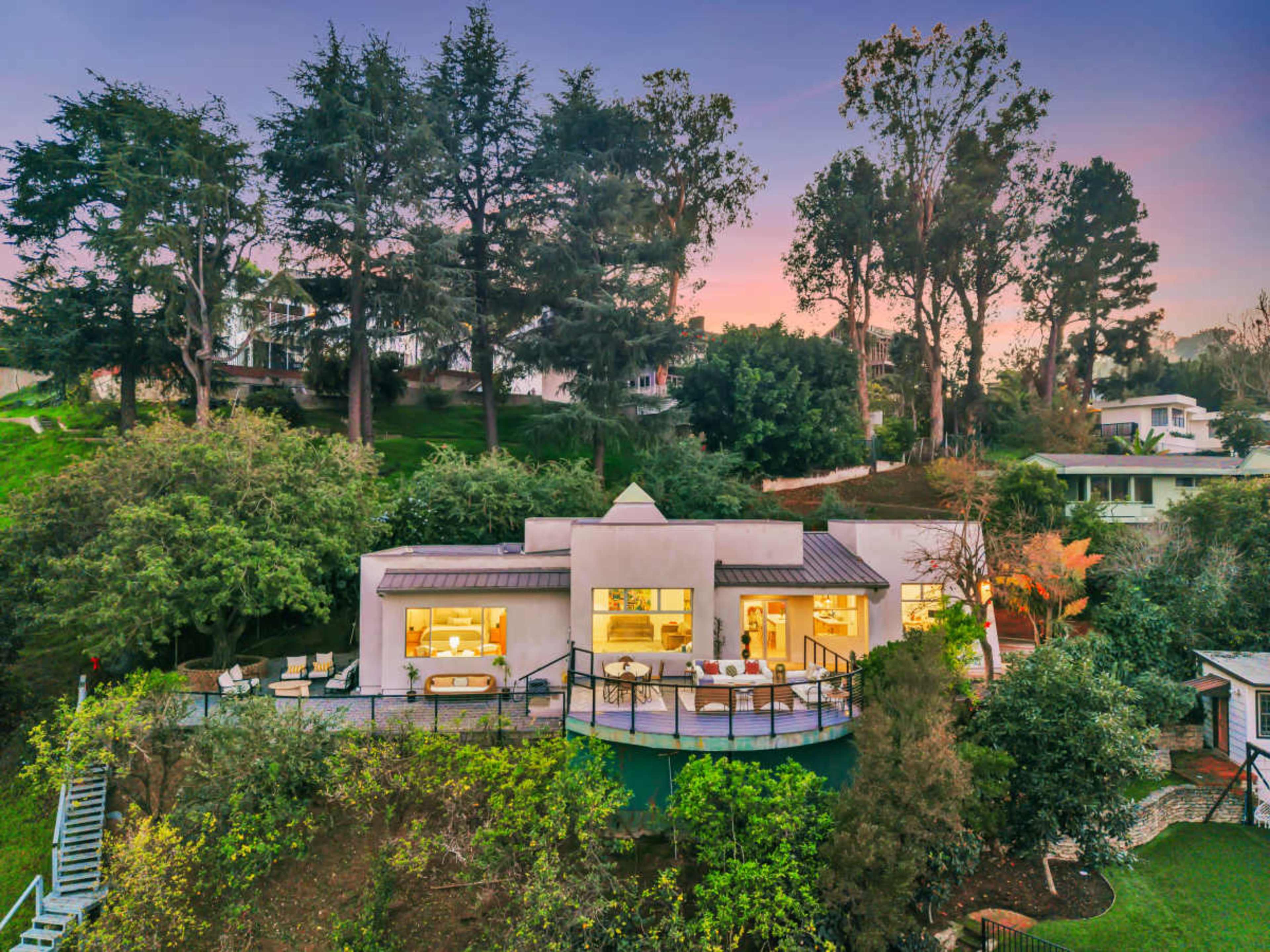 A modern house with large windows and a deck is situated on a hillside surrounded by trees and other homes.