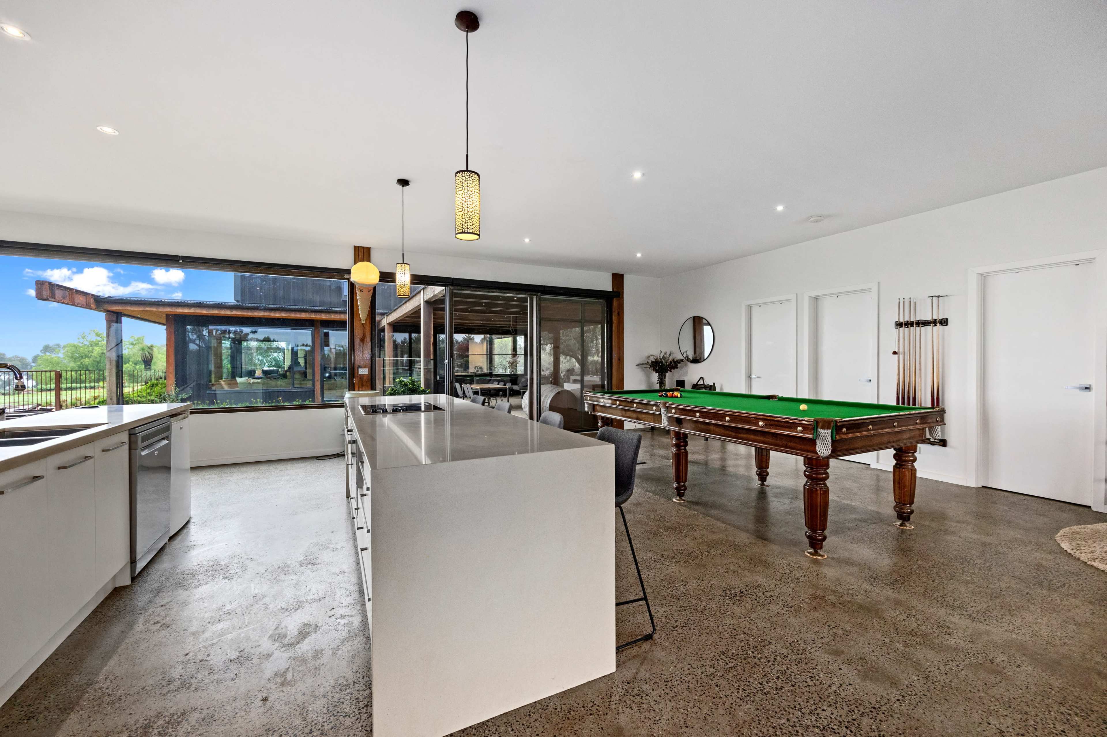 A modern open-concept room with a kitchen area and a billiard table, showcasing large windows that provide a view of the outdoors.