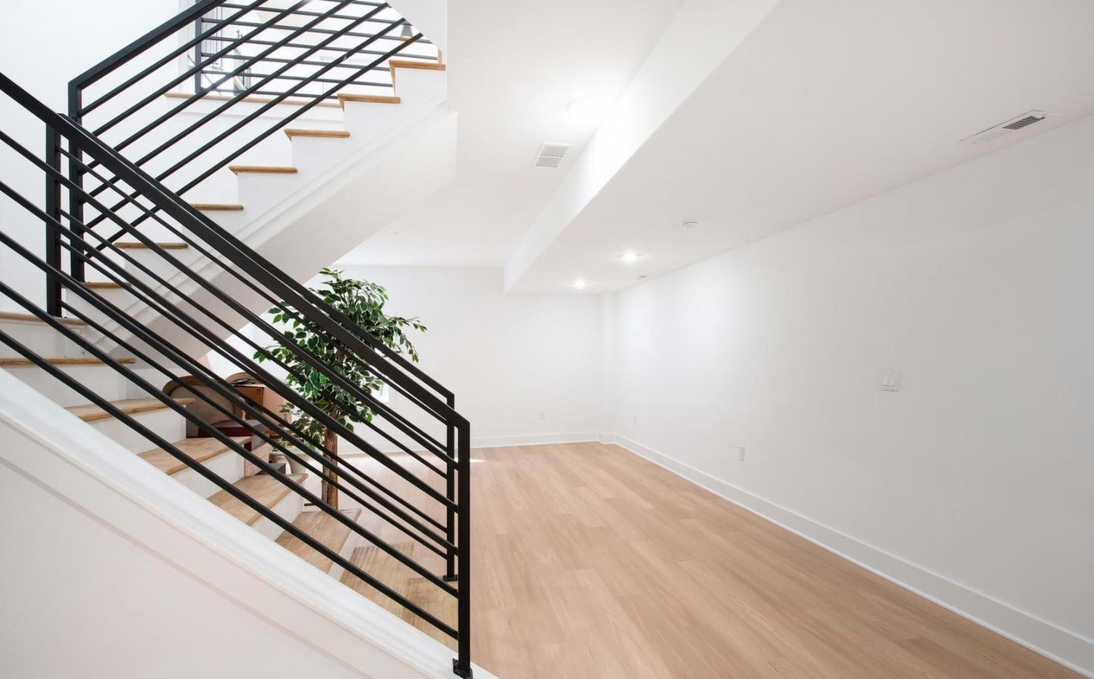 A staircase with a black railing ascends to a bright, minimalistic room with light wood floors and a small potted plant in the corner.