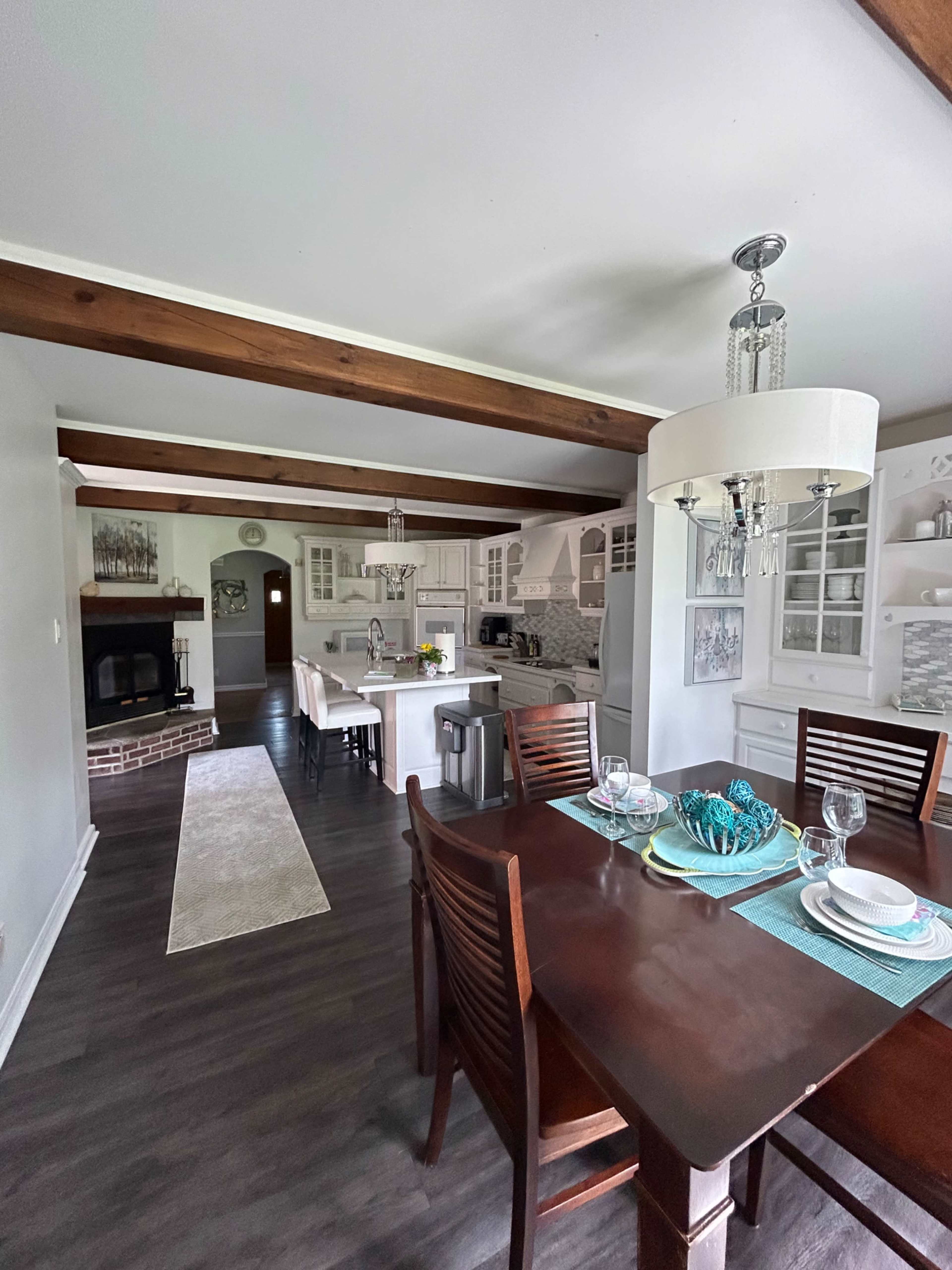 A modern kitchen and dining area with wooden beams, a dining table set for four, and a cozy fireplace in the background.