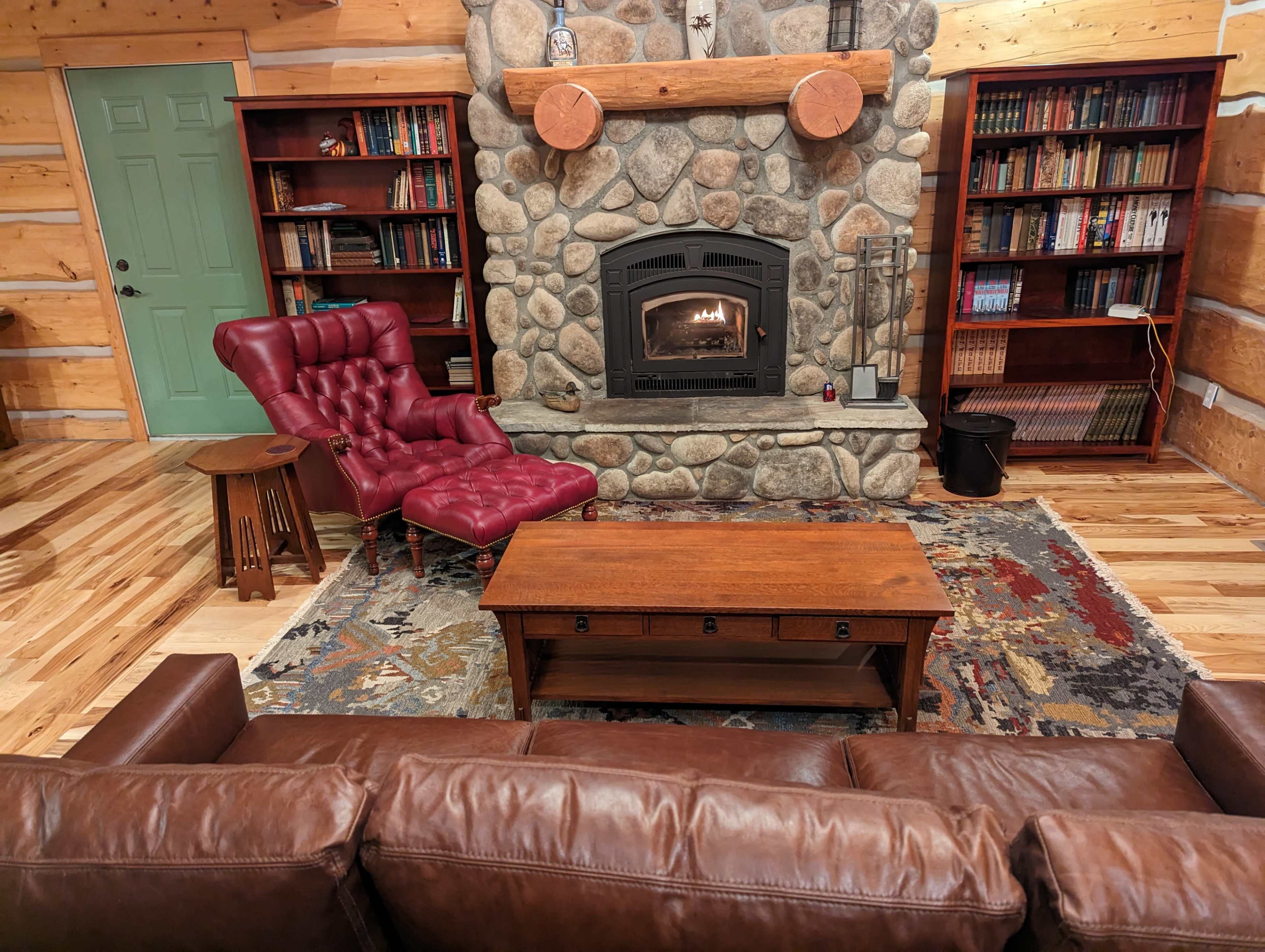 A living room features a stone fireplace with a fire burning, surrounded by a red armchair, a brown couch, a wooden coffee table, and bookshelves filled with books.