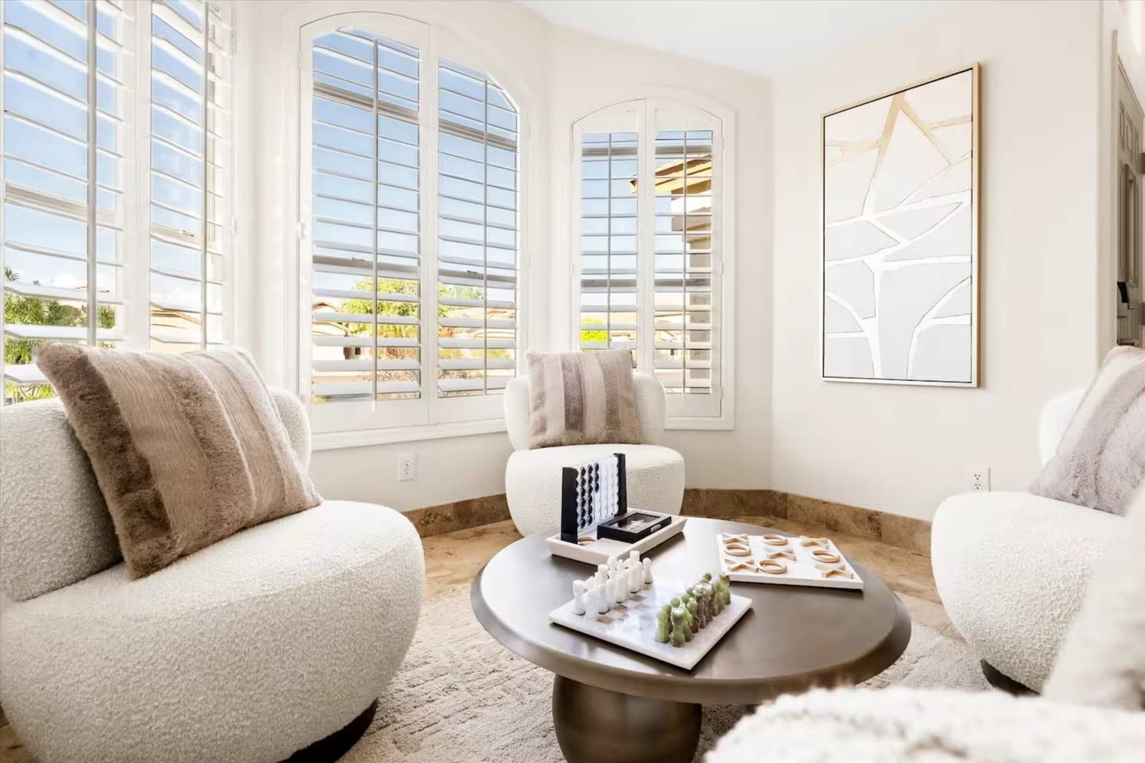 The image depicts a bright, cozy seating area featuring two upholstered chairs and a round coffee table, with large windows allowing natural light to flow in.