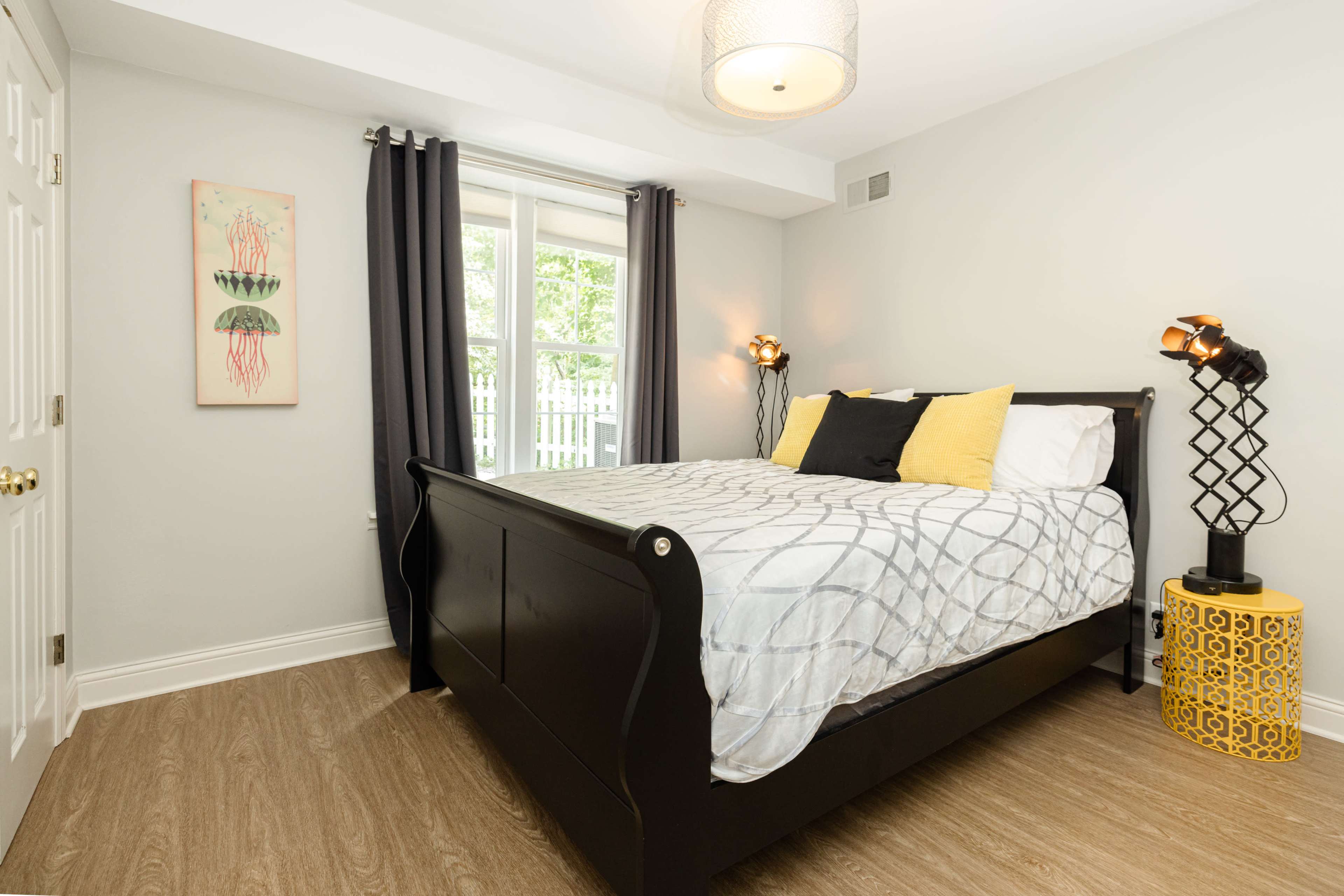 A modern bedroom features a black sleigh bed with gray and white bedding, two side lamps, and a yellow accent table by a window with sheer curtains.