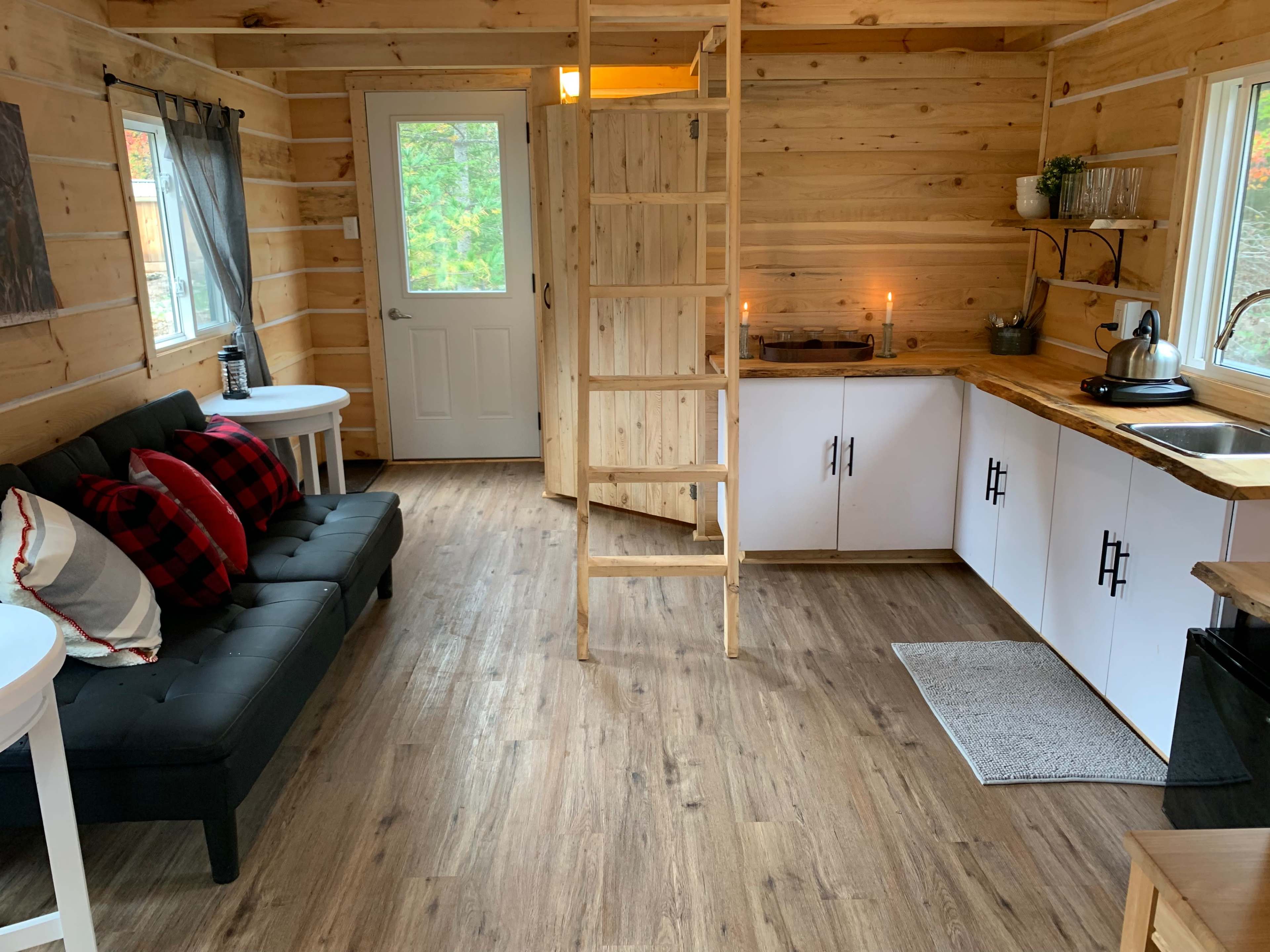 The image shows a cozy, wooden interior of a small cabin with a ladder leading to a loft, a black futon, and a kitchenette.