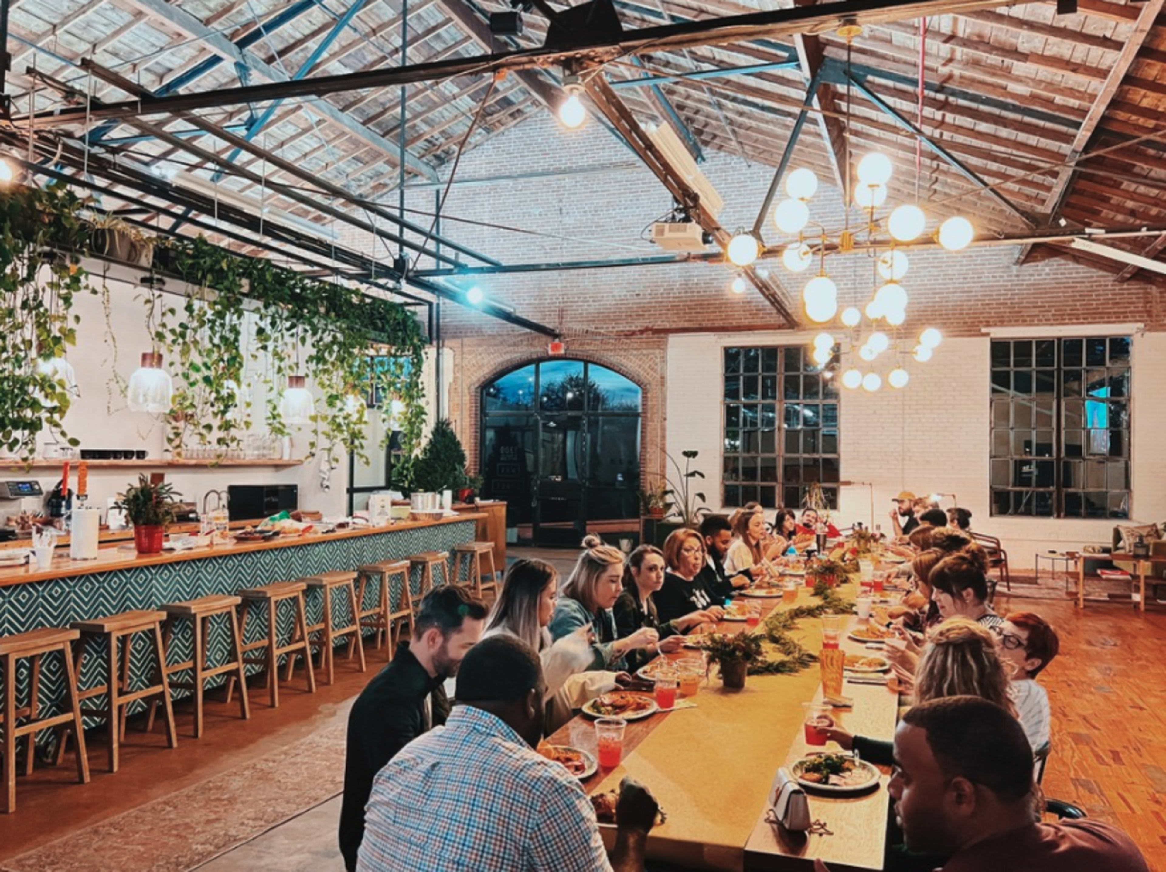 A group of people gathers around a long table arranged for a meal in a spacious, well-lit dining area with exposed brick walls and hanging plants.