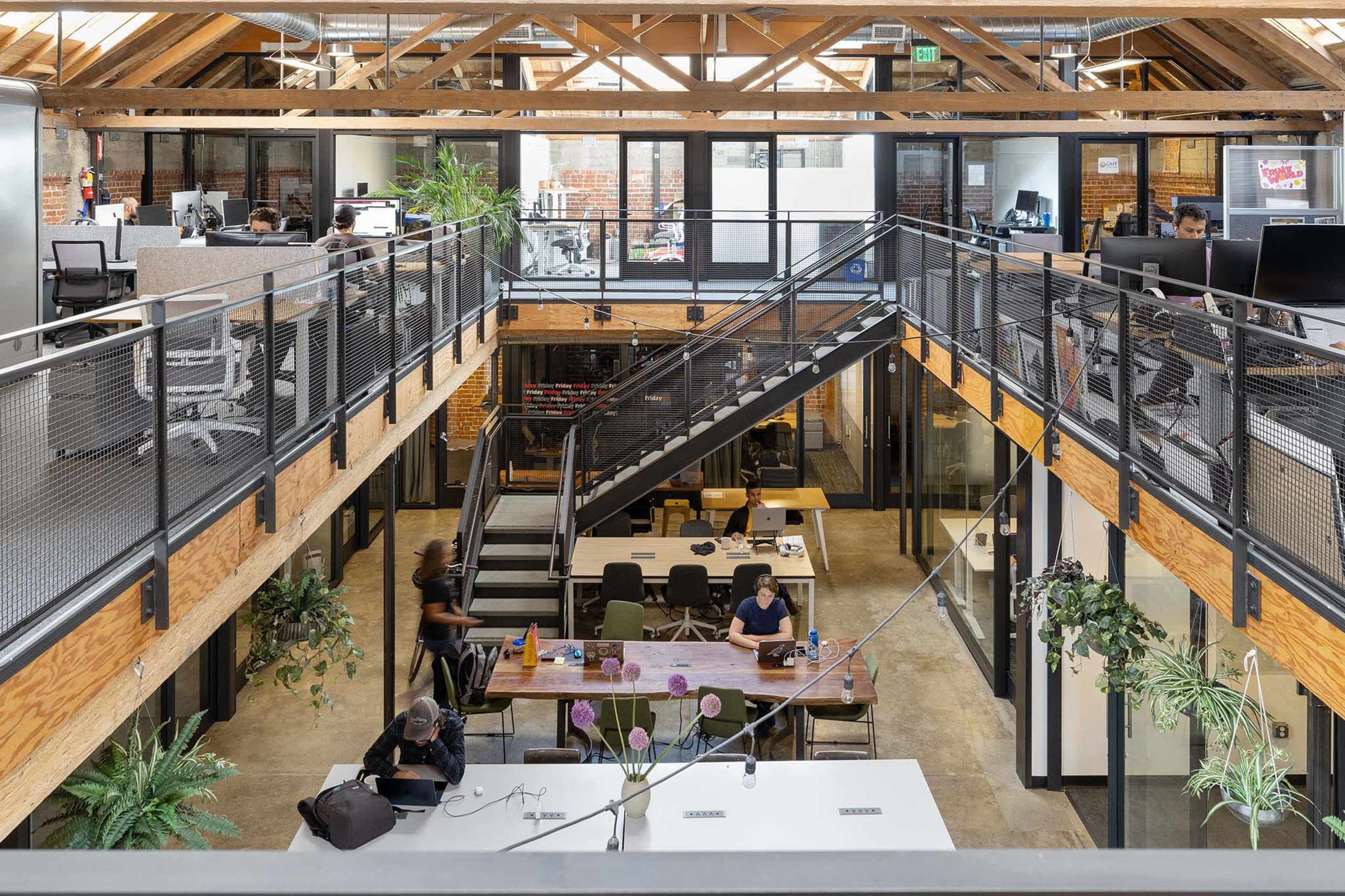 An open-plan office space with multiple workstations, a central staircase, and large windows that allow natural light to enter.