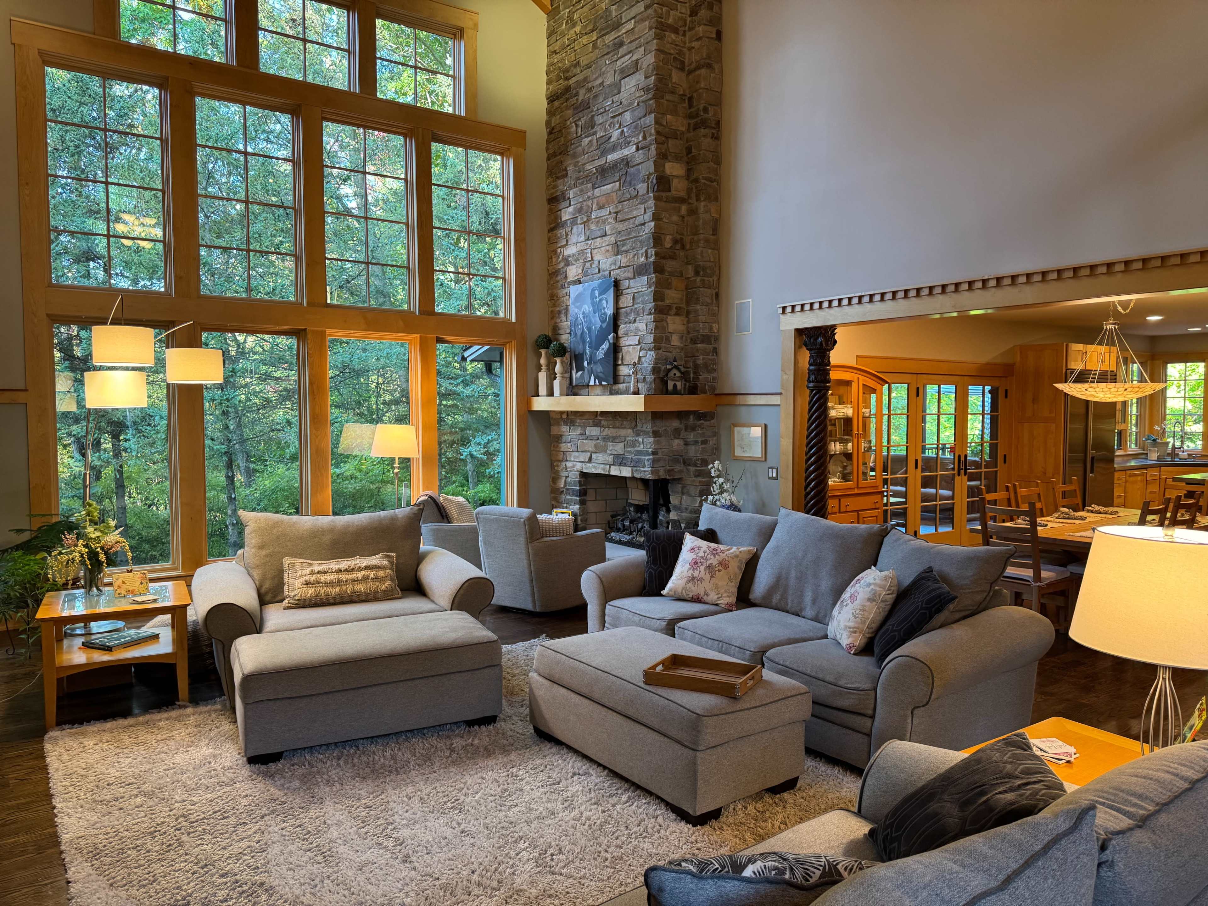 The image shows a spacious living room with large windows, a stone fireplace, and comfortable furniture arranged on a soft rug.