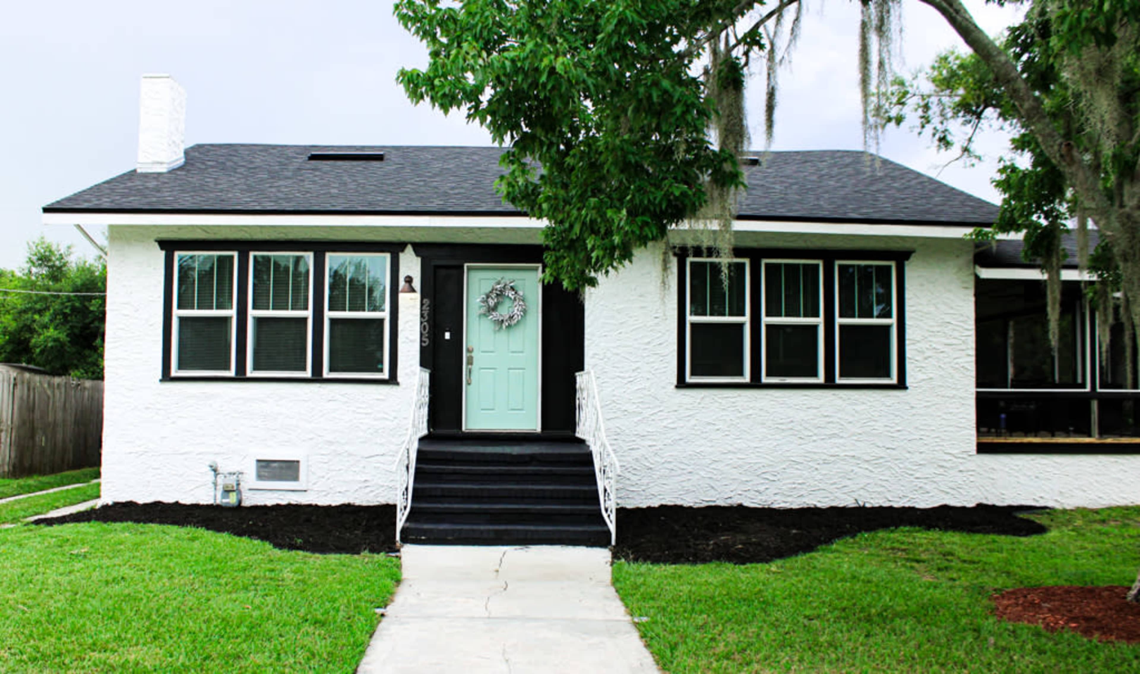 The image shows a single-story white house with a black roof, green front door, and a landscaped yard featuring grass and shrubs.