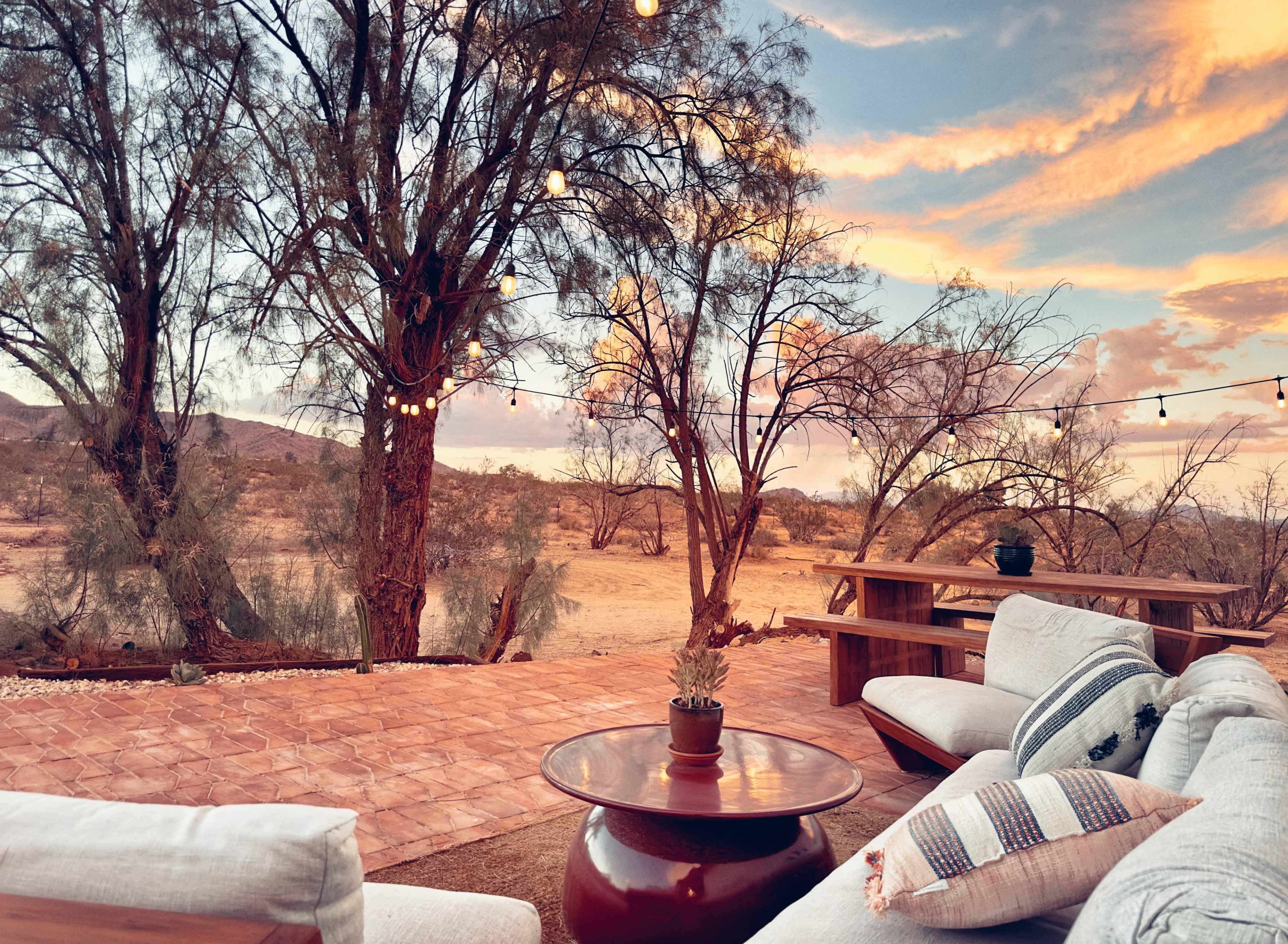 A patio area features comfortable seating and a wooden table overlooking a desert landscape with trees and a colorful sunset sky.