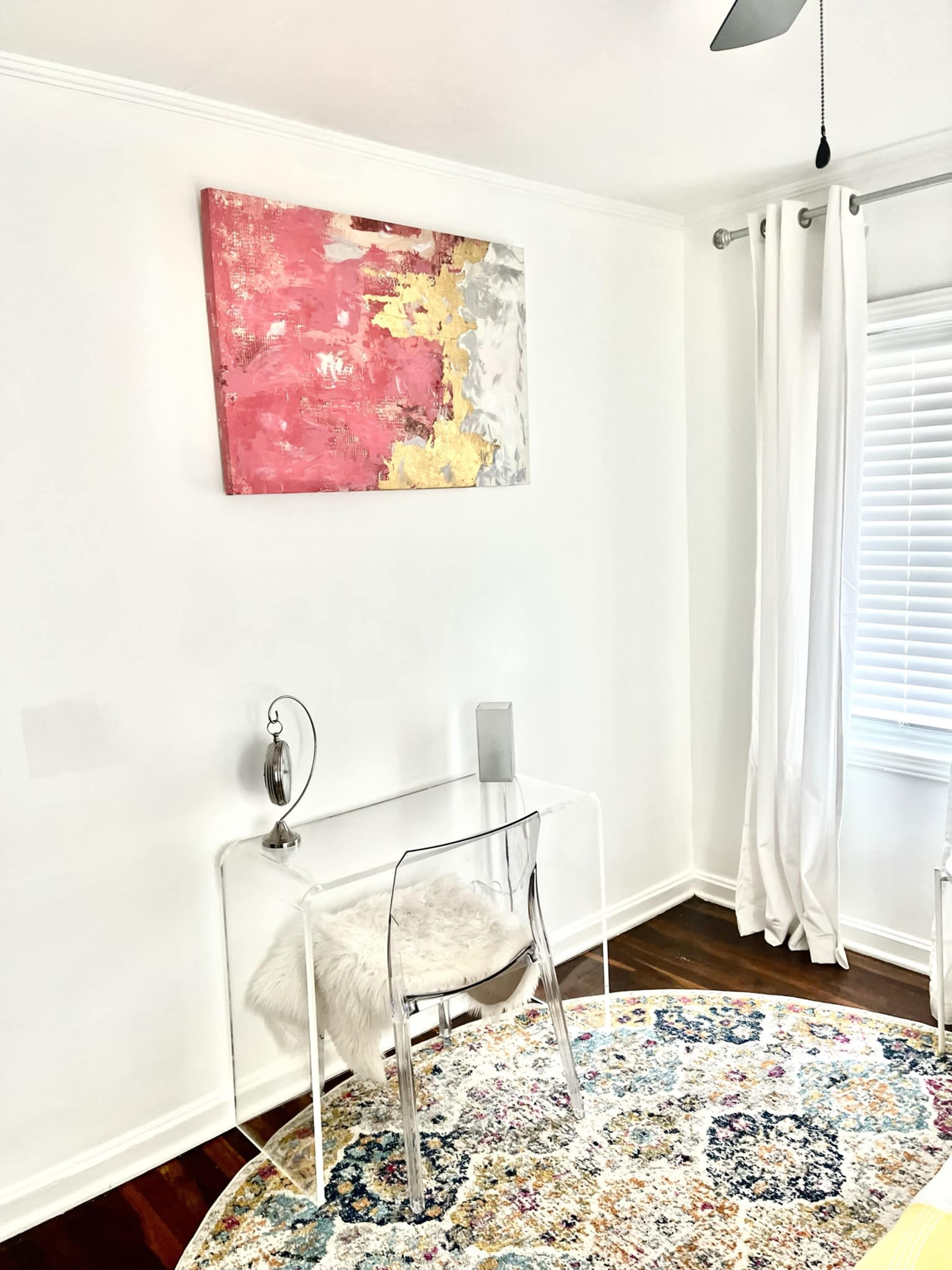 A small, minimalist workspace featuring a clear acrylic chair and a simple glass table set against a white wall, with a colorful abstract painting above and a round patterned rug below.