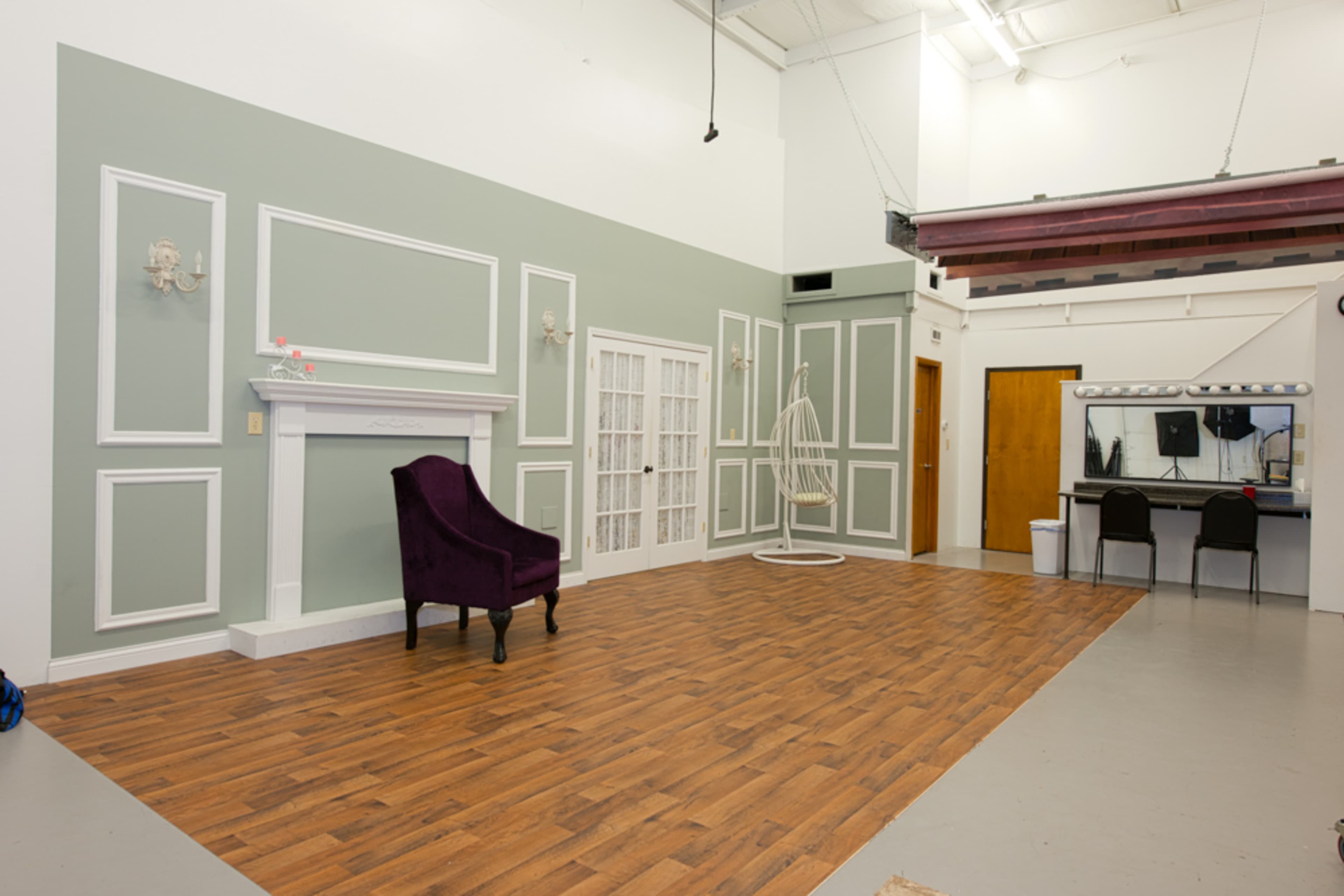 A spacious studio featuring a hardwood floor, a decorative fireplace, a purple chair, and mirrors along with a styling station against a light green wall.