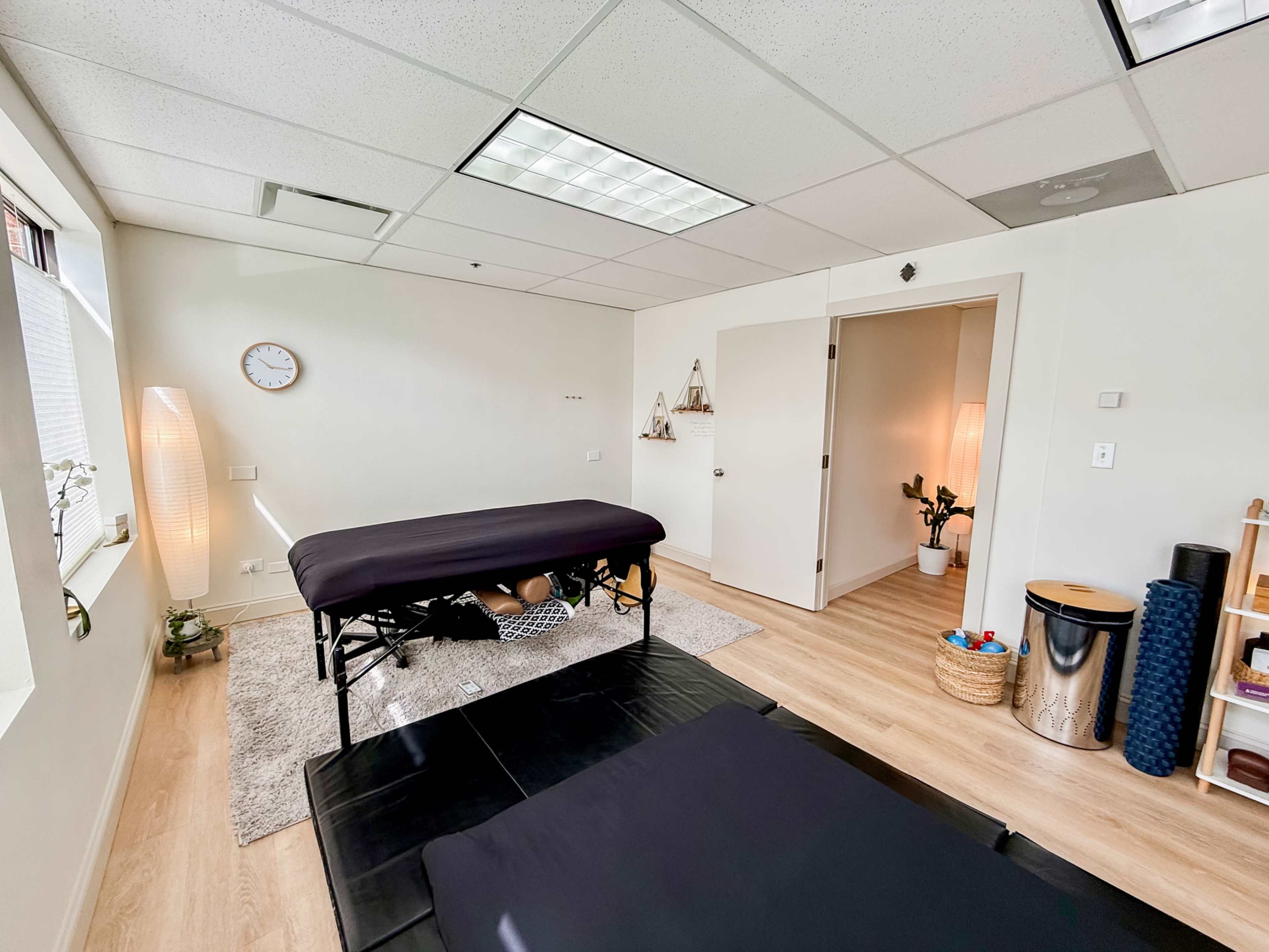 The image shows a massage therapy room with two treatment tables, a wall clock, and a small plant in the corner.