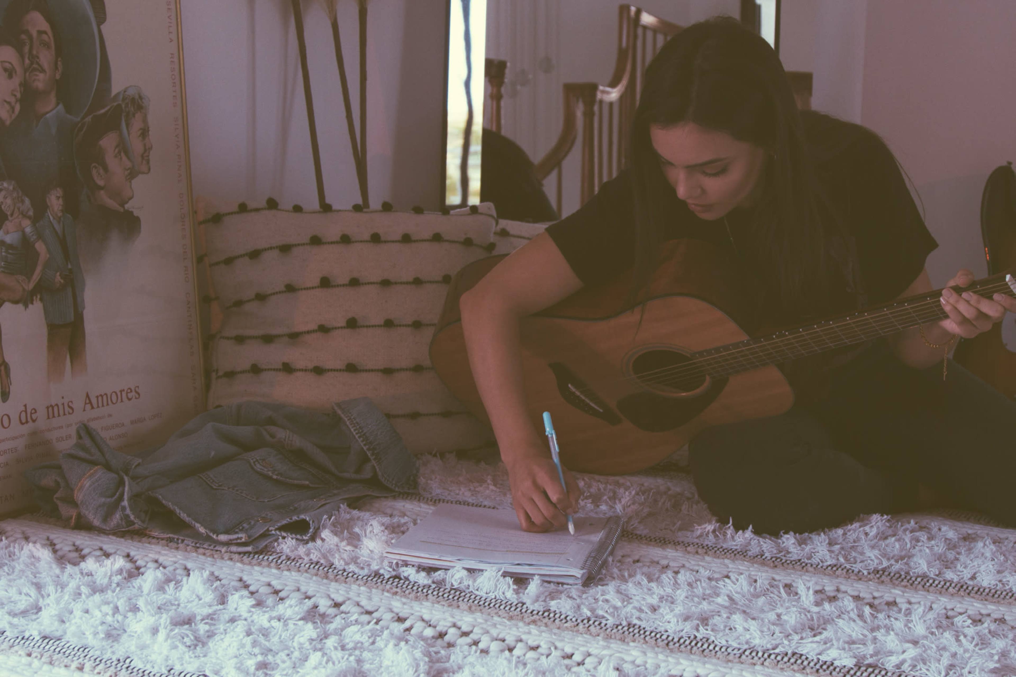 A person sits on a rug with a guitar in their lap while writing in a notebook.