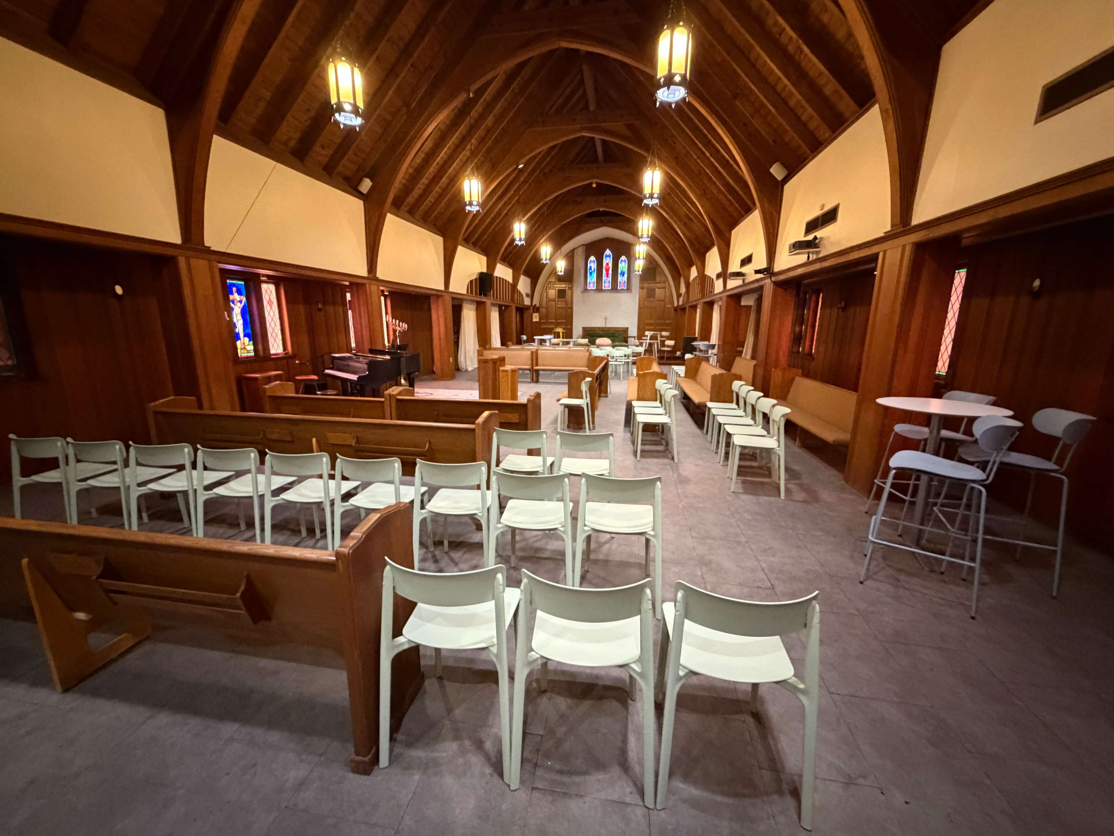 The interior of a wooden church features rows of pews and chairs, with stained glass windows and a high, arched ceiling.