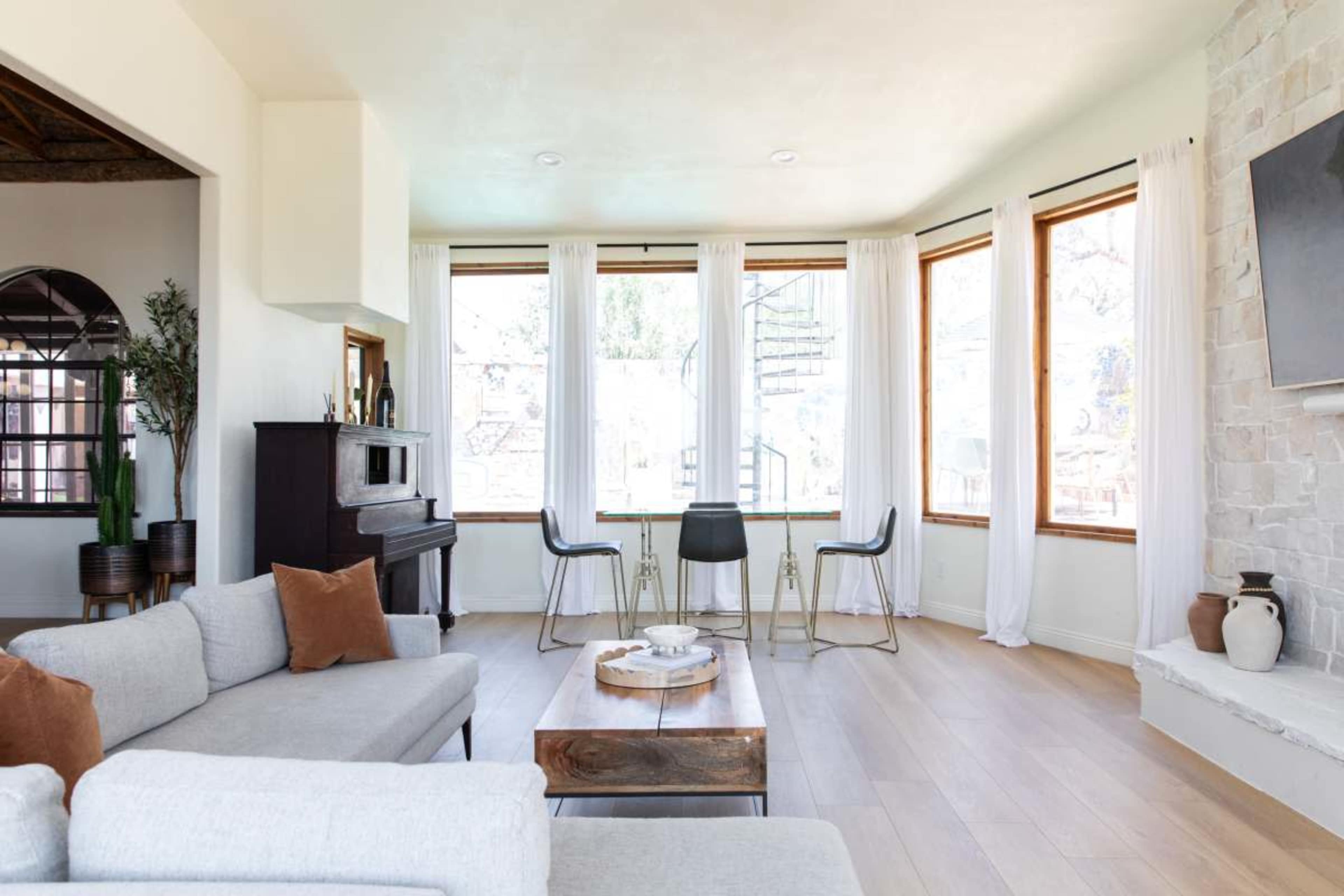 A light-filled living room features a gray couch, a wooden coffee table, and three black chairs around a windowed area.