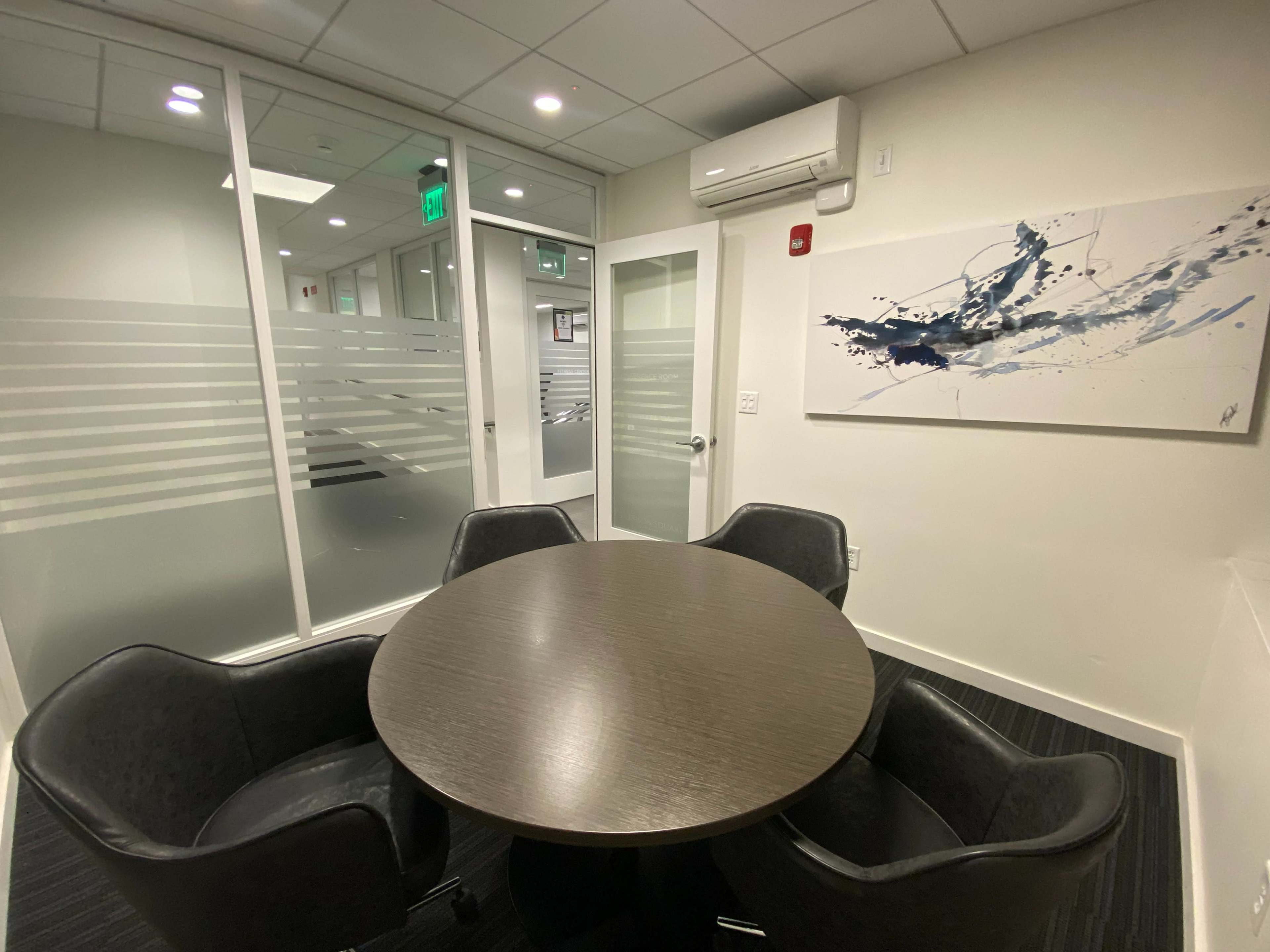 A small conference room features a round table surrounded by four black chairs, with glass walls and an abstract painting on one side.