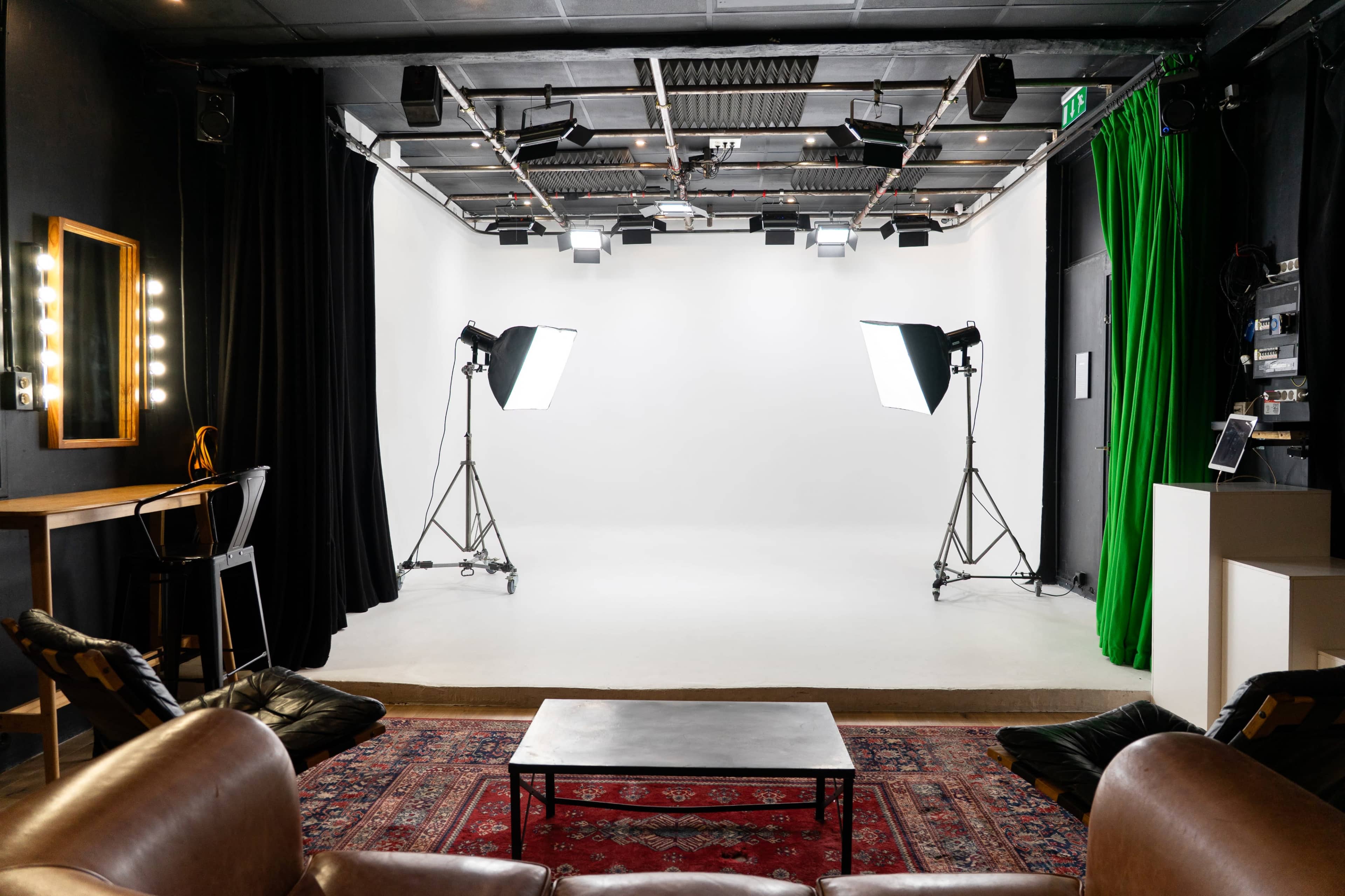 The image shows a photography studio with a white backdrop, two lighting setups on tripods, and a seating area featuring a coffee table and a patterned rug.
