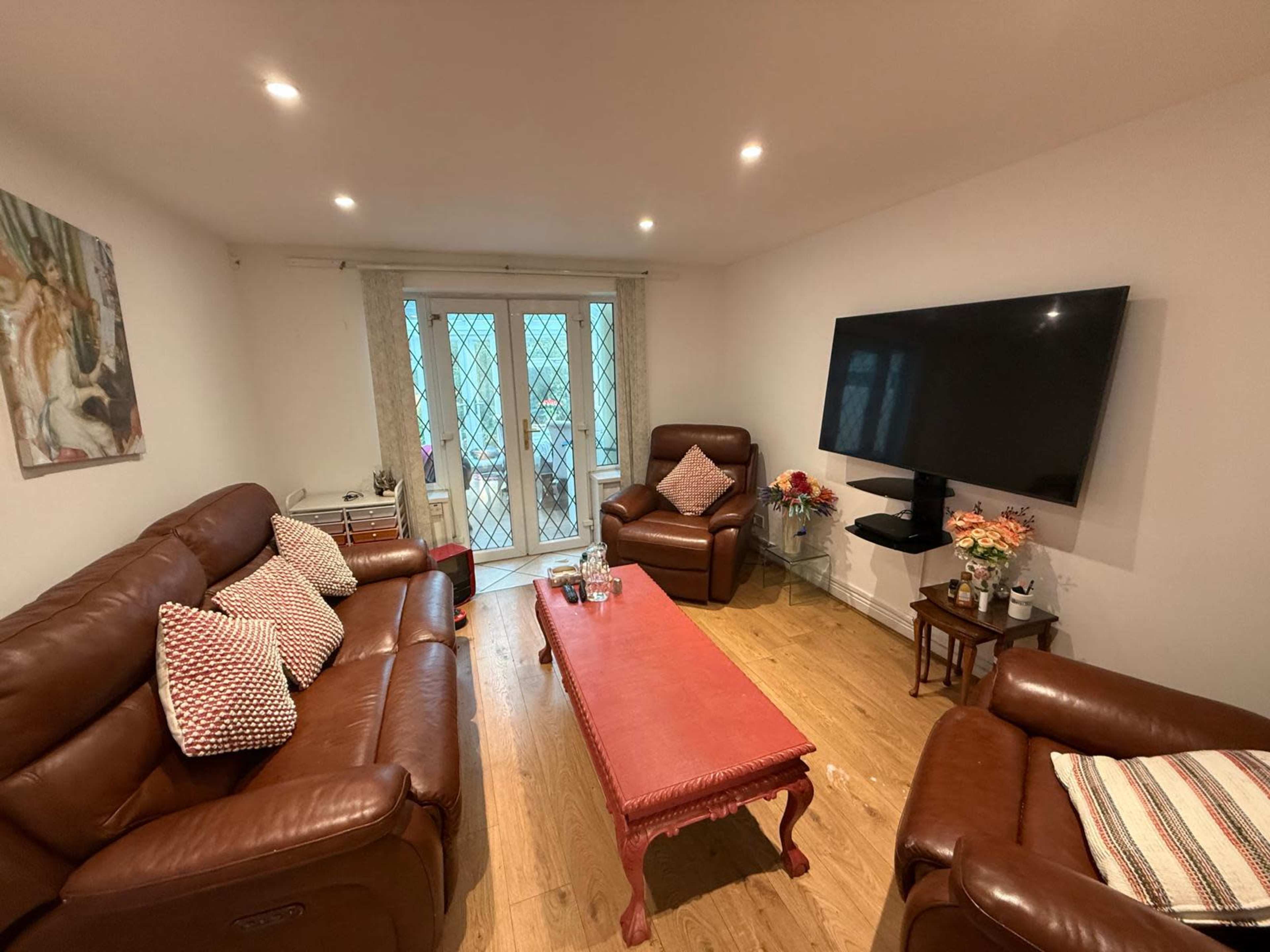 The image shows a living room with two brown leather sofas, a red coffee table in the center, a television mounted on the wall, and sliding glass doors leading to an outdoor area.
