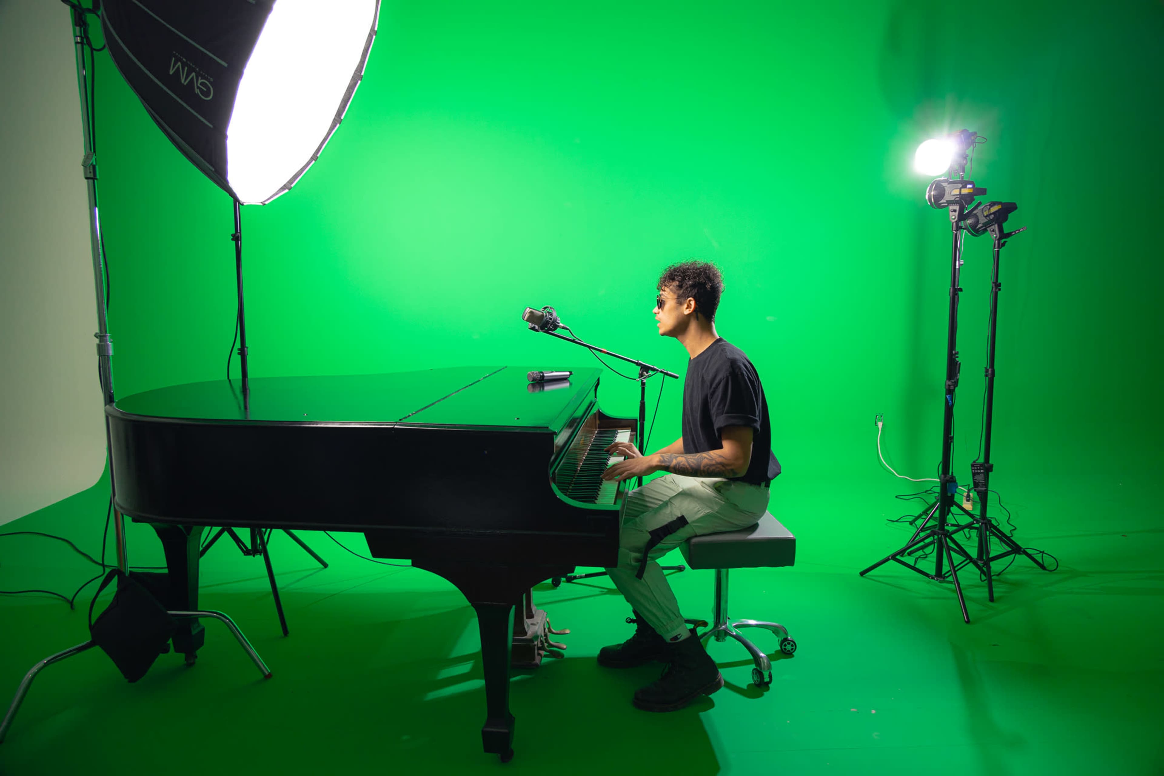 A person sits at a grand piano in front of a green screen, with studio lights positioned nearby.