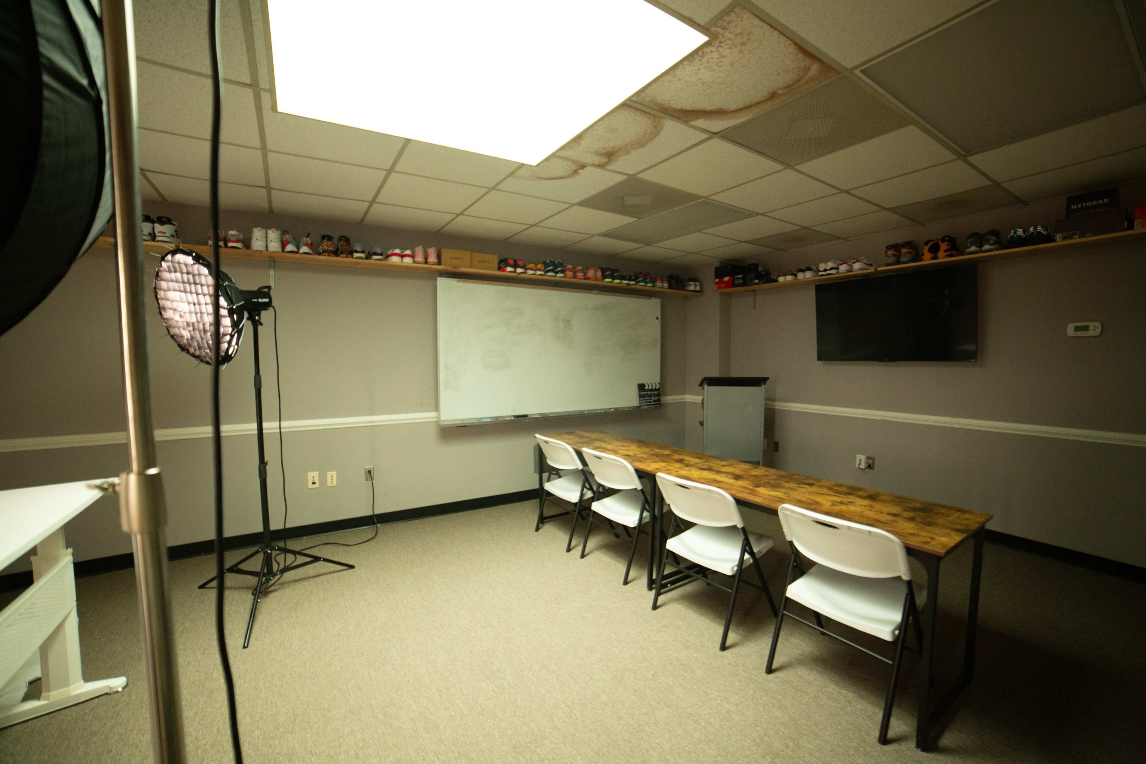 A minimal conference room features a long wooden table, six white chairs, a large whiteboard, and a television mounted on the wall, with shelves above displaying various shoe boxes.
