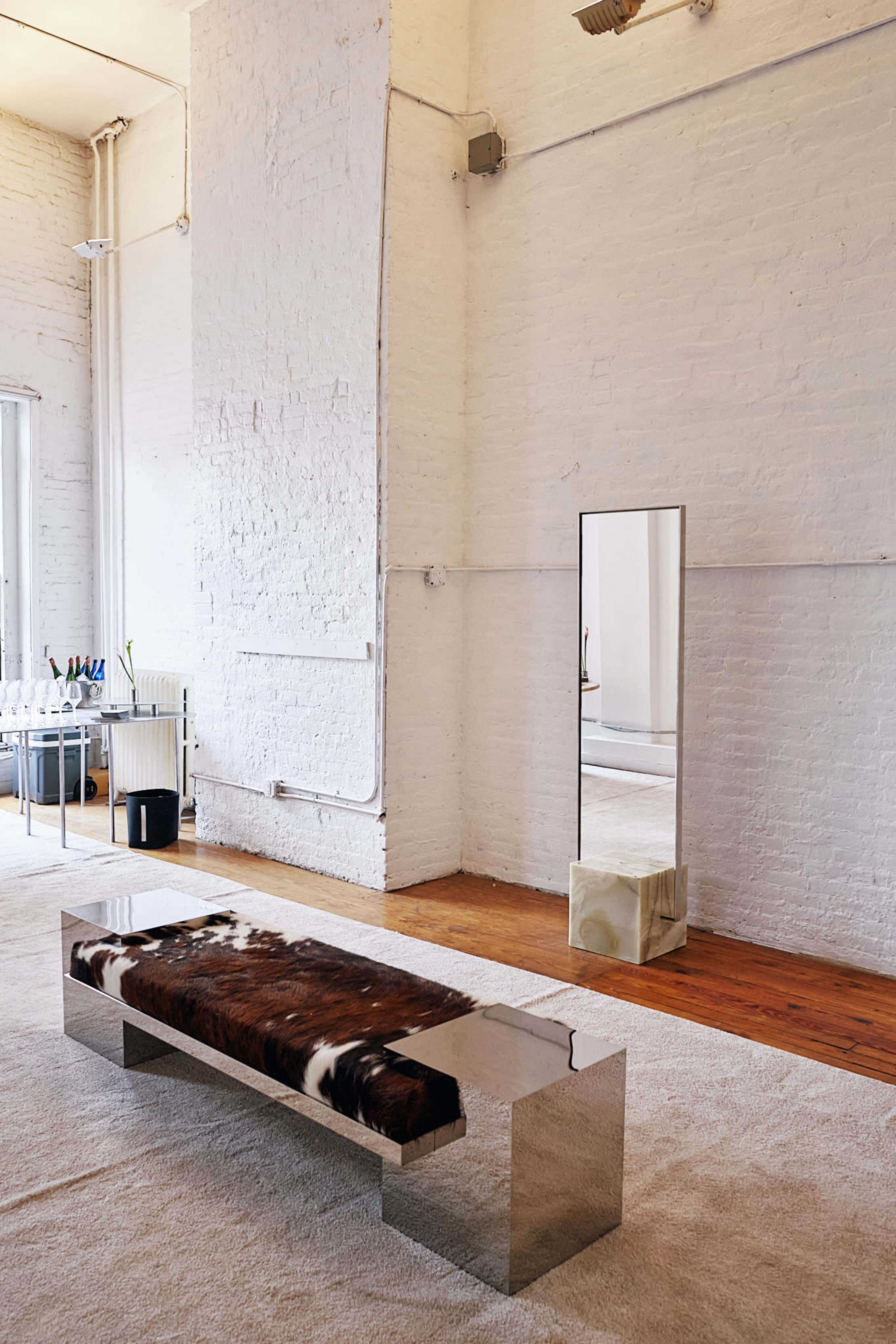 The image shows a minimalist interior featuring a modern, mirrored bench with a cowhide cushion, a large wall-mounted mirror, and a light-colored area rug.