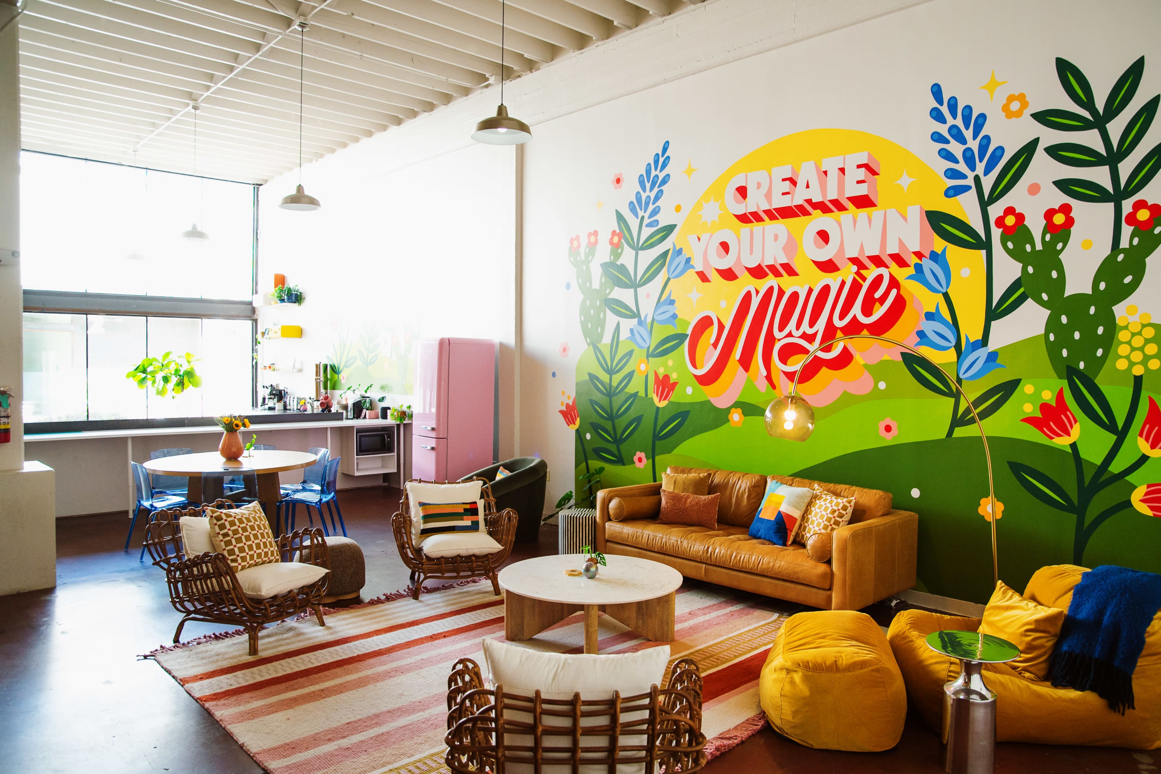 The image shows a bright, colorful lounge area with a large mural featuring the phrase "Create Your Own Magic," surrounded by plants and decorative furniture.