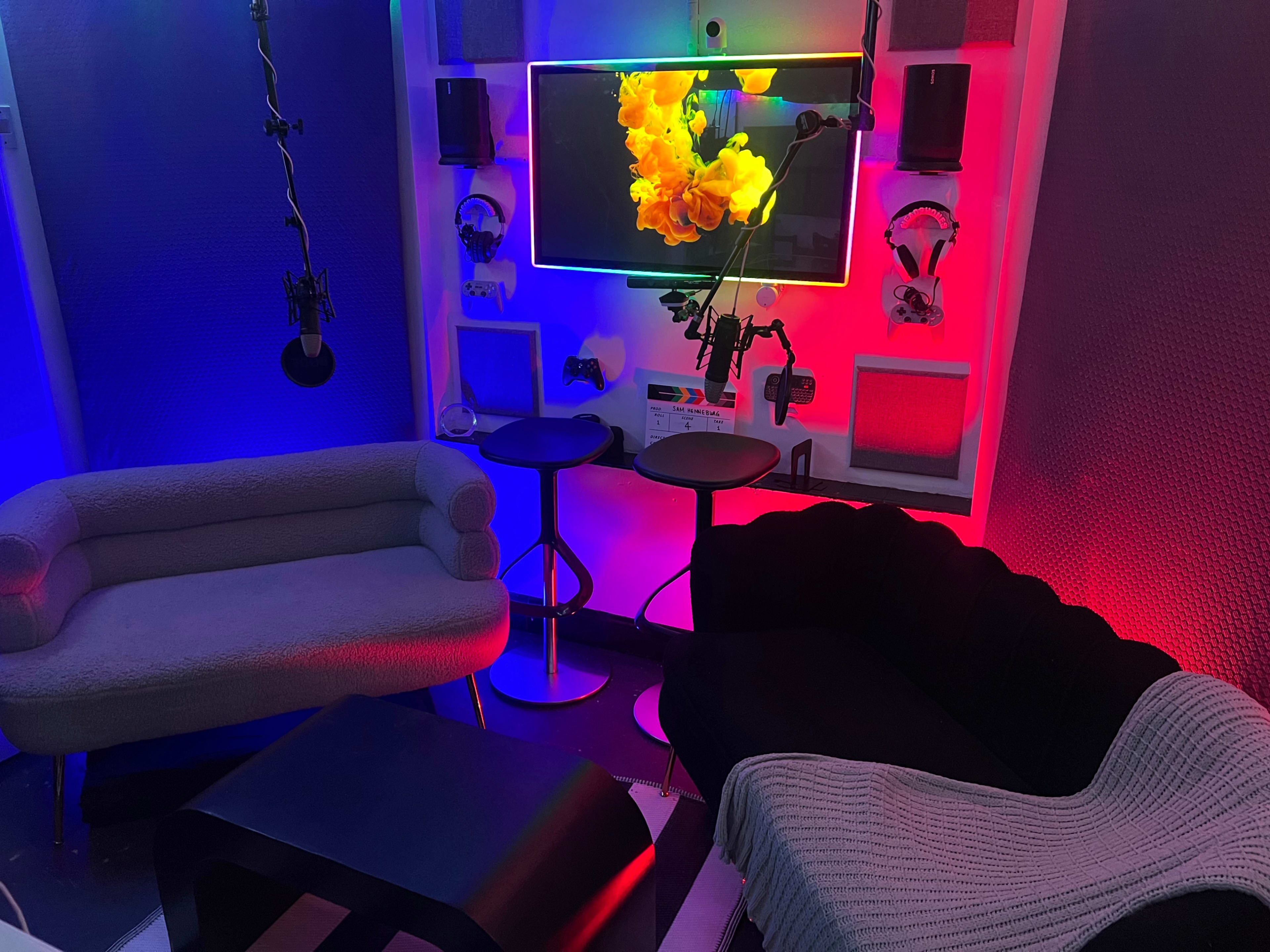 The image shows a modern, colorful lounge area with a television mounted on the wall, surrounded by stylish furniture including two sofas and bar stools.