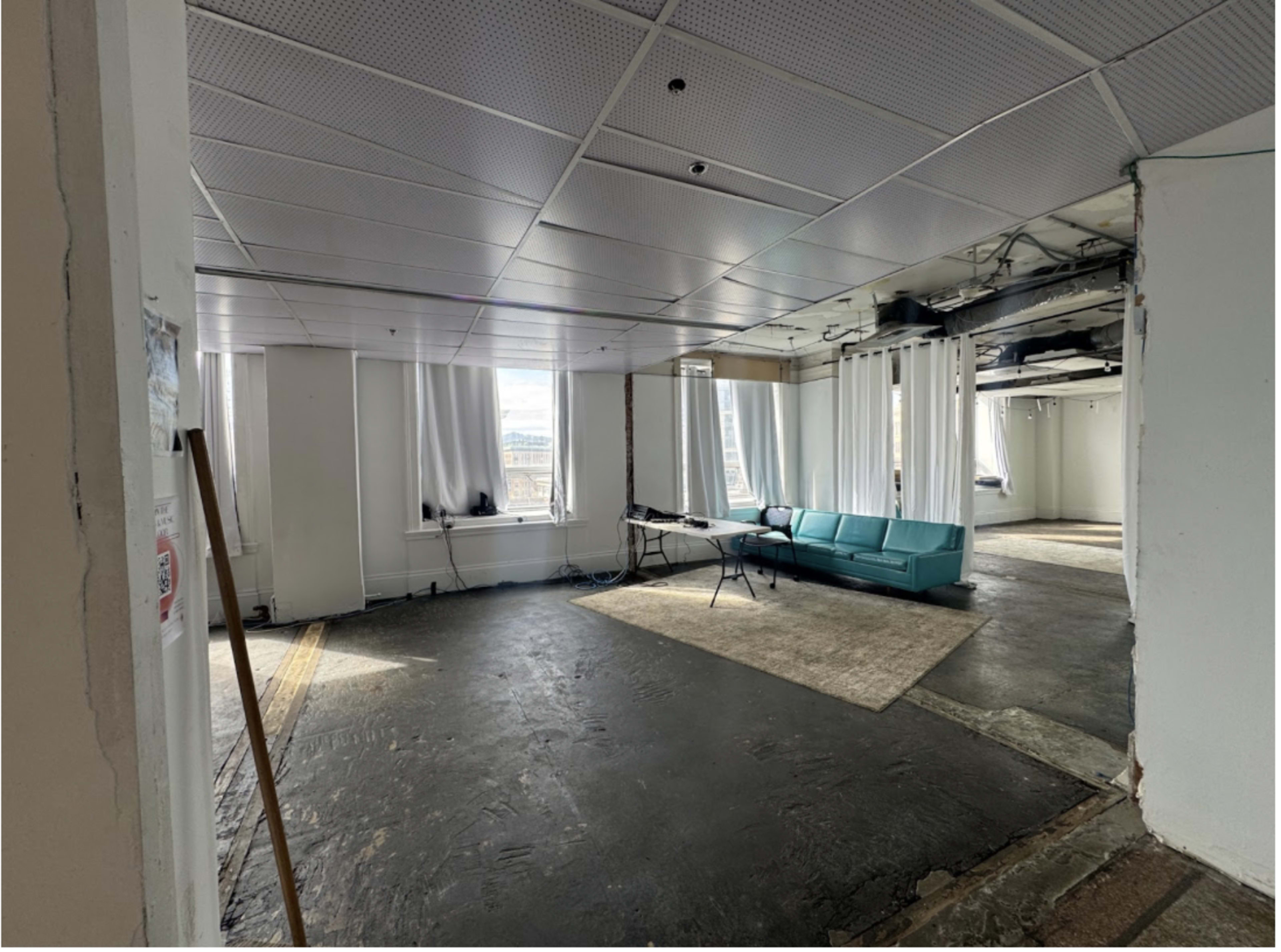 The image shows a large, vacant room with a teal couch, a table, and windows covered by white curtains.