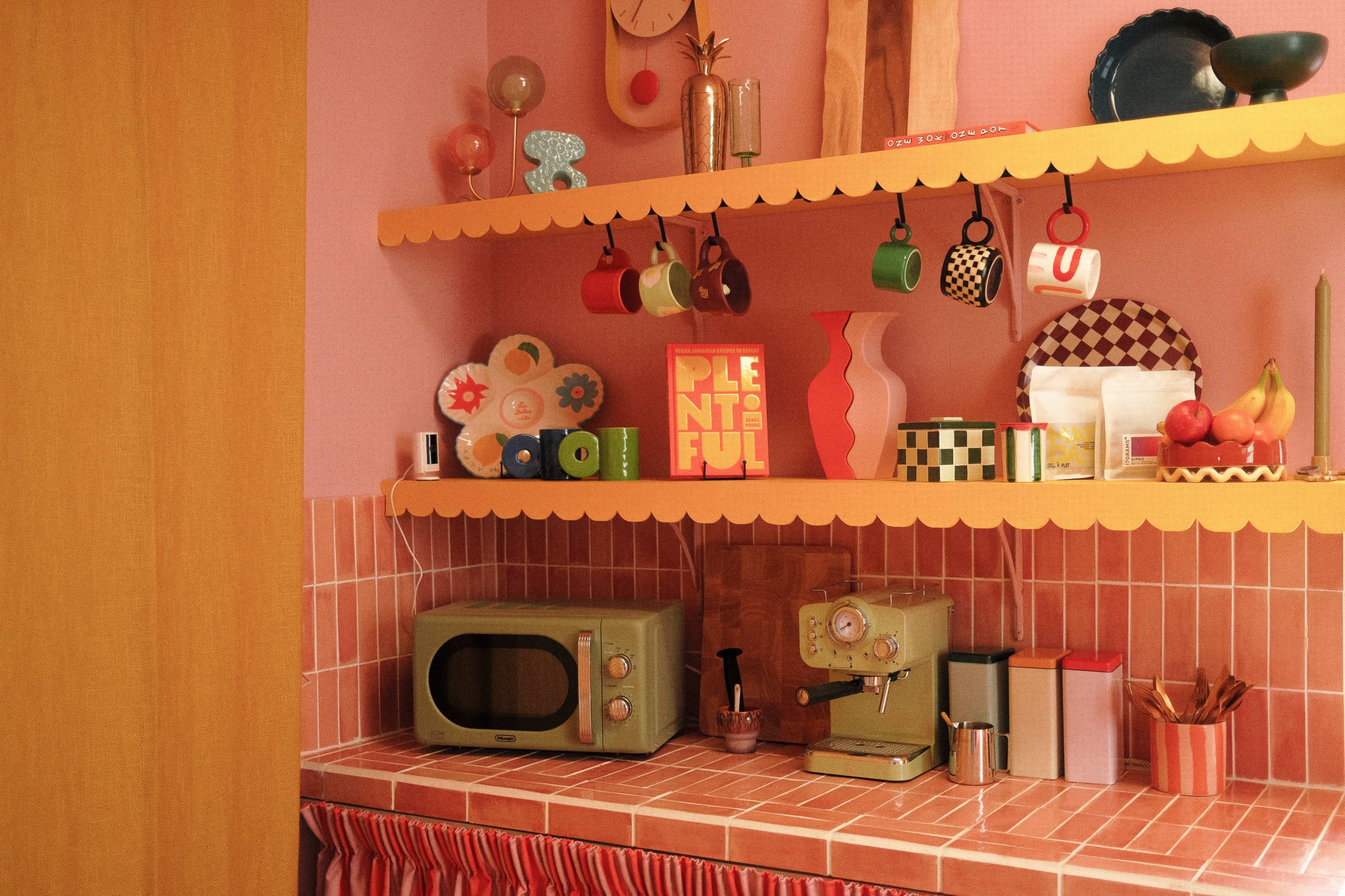 The image shows a vintage-style kitchen with pink walls, a checkered countertop, and colorful dishware arranged on shelves.