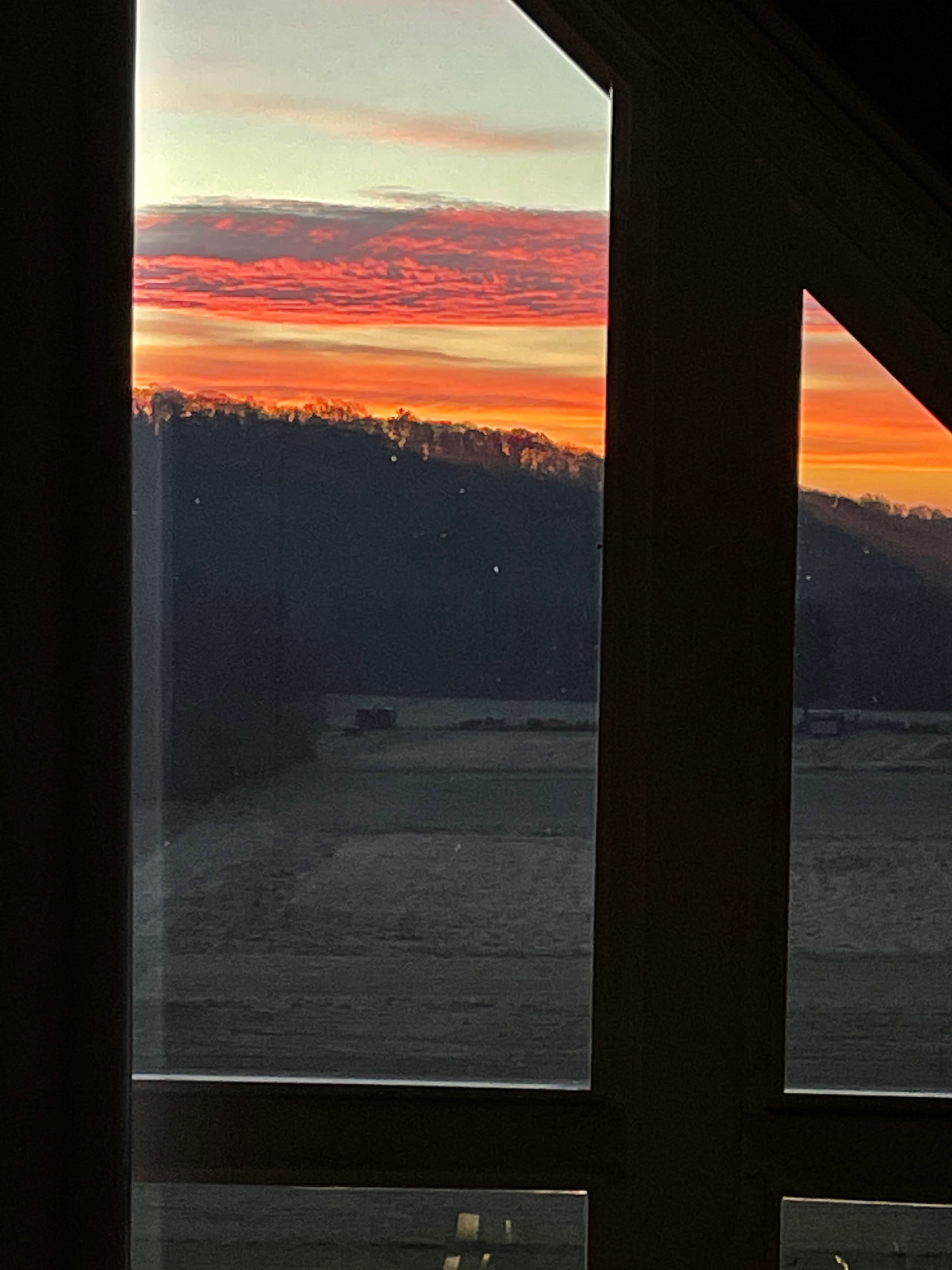 A sunset with vibrant clouds over a tree-lined horizon, viewed through a large window.
